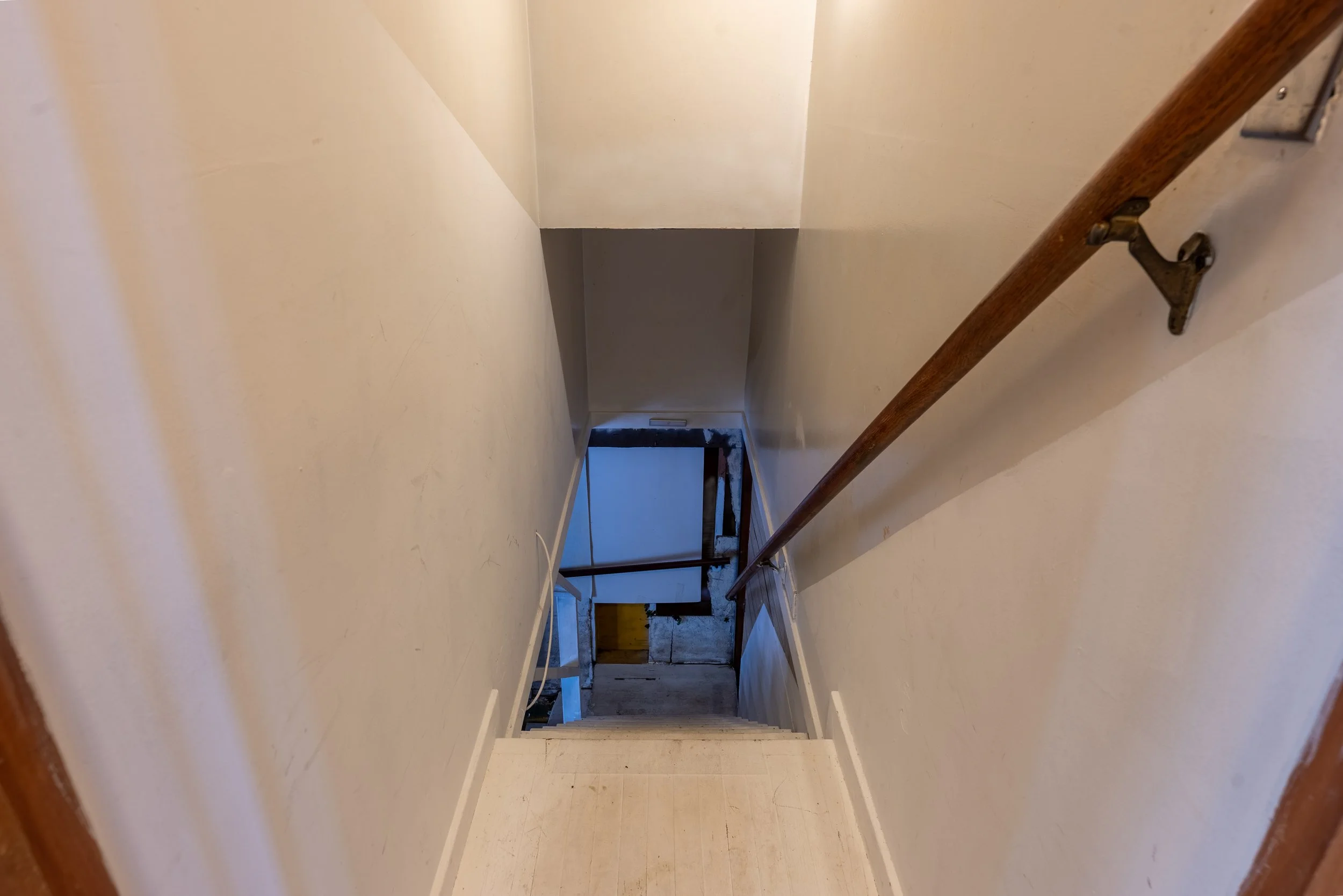 Empty hallway or staircase area with beige walls, brown handrail, and visible doorway at the end.