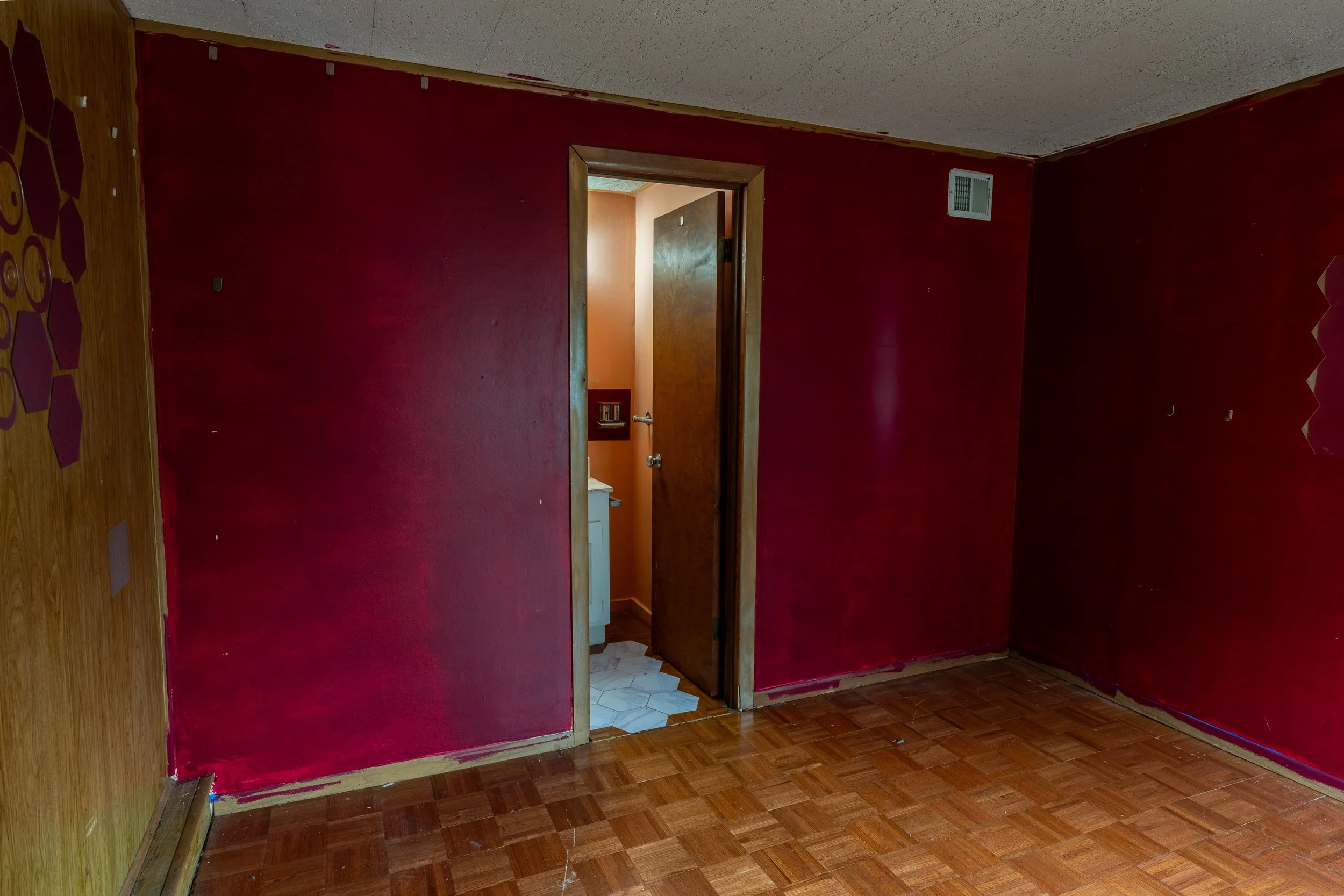 An empty room with red walls, partially unfinished, with a doorway leading to a bathroom with orange walls and white tile flooring.