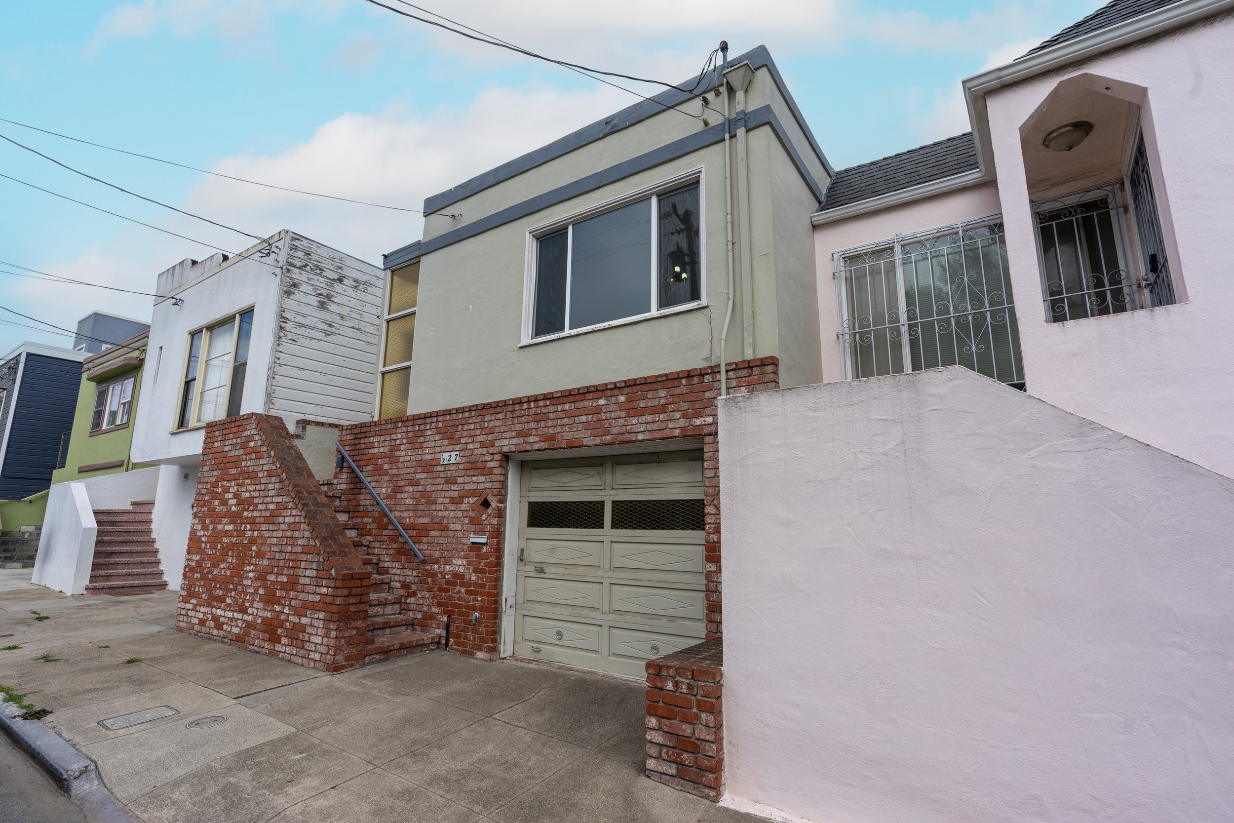 A row of three houses with stairs leading up to their entrances, painted in different colors: green, white, and pink. The houses have various window styles, with some barred windows. The sky is partly cloudy, and power lines are overhead.