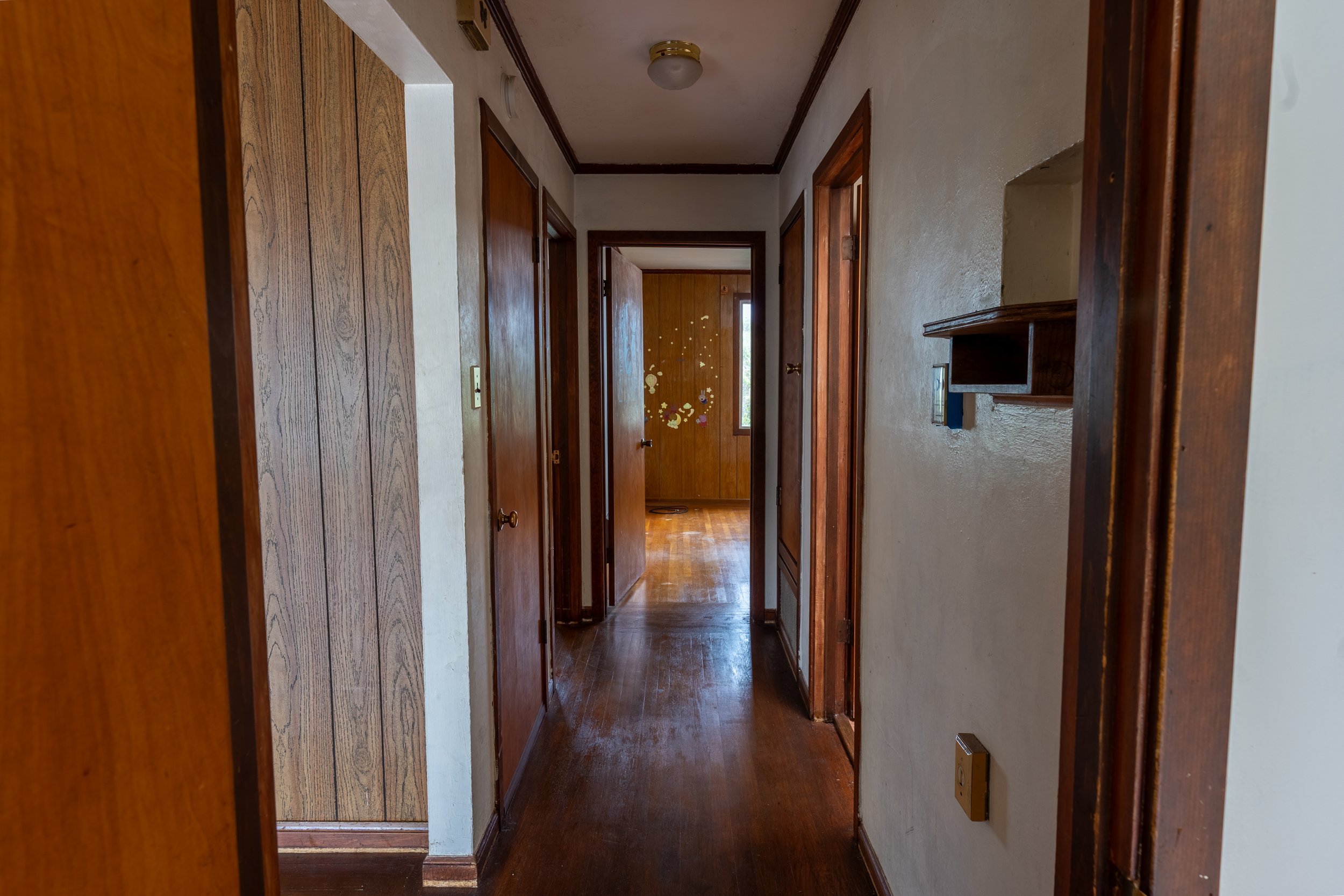 A hallway in a house with wood-paneled walls and hardwood flooring, leading to a door decorated with stickers. There is a window at the end of the hallway and a ceiling light fixture.