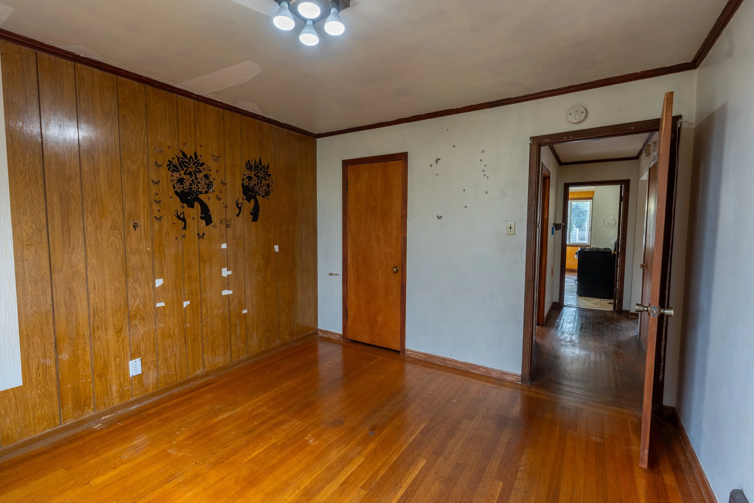 Empty room with wooden wall paneling, hardwood floors, and a ceiling light fixture. There is a doorway leading to another room with visible window and door. The wall has some small decorative stickers and marks.