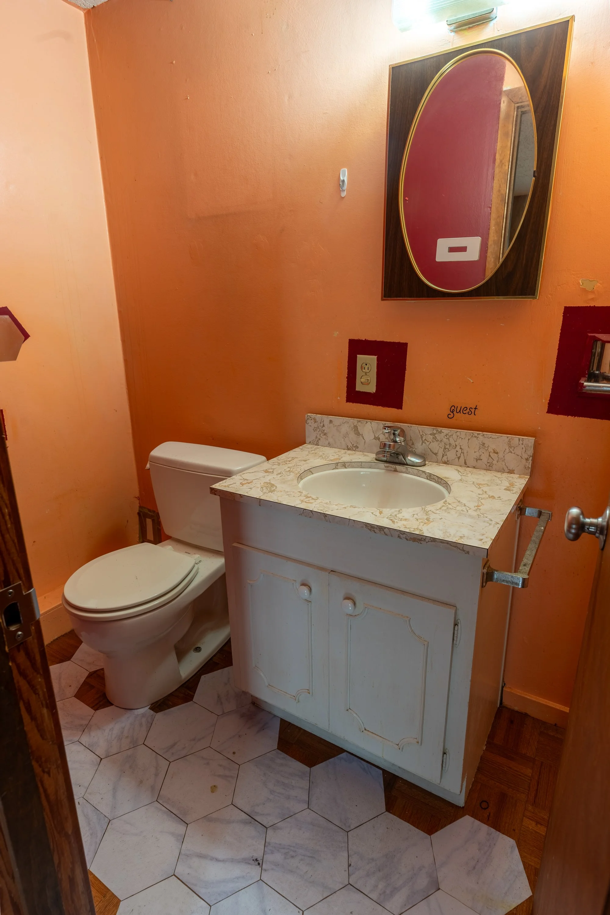 A small bathroom with orange walls, a white toilet, a white cabinet with a granite top and oval sink, a mirror with a wood frame, and a wall vent light above the mirror. The floor is a mix of marble and brown wooden tiles.
