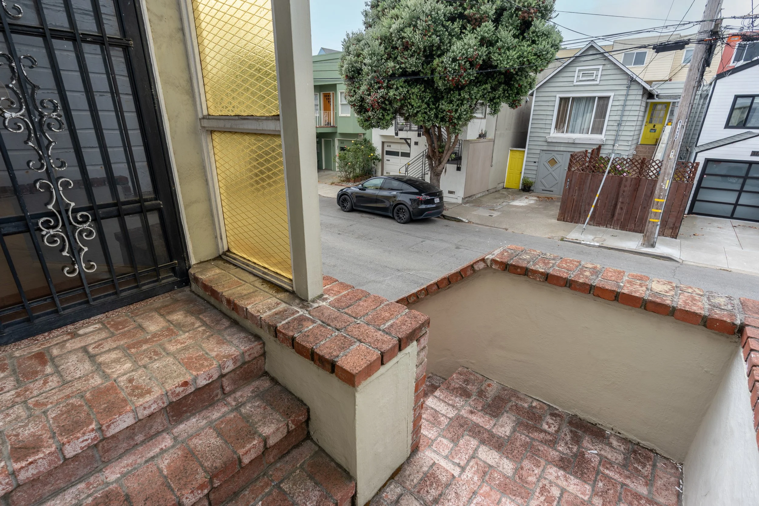 View from a porch or balcony looking out onto a residential street with trees, cars, and houses, including a black car parked on the street and colorful houses with yellow and gray doors.