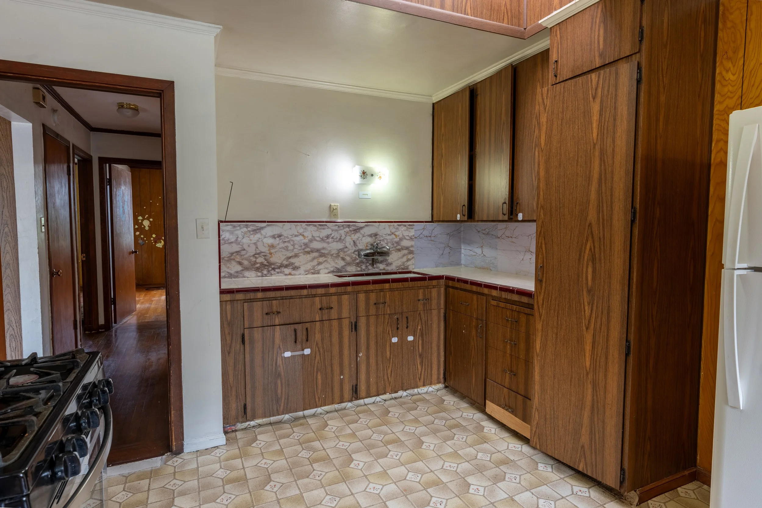 A kitchen with wooden cabinets, a marble backsplash, and a gas stove in the lower left corner. The floor has a patterned linoleum covering, and a hallway with wooden doors is visible in the background.