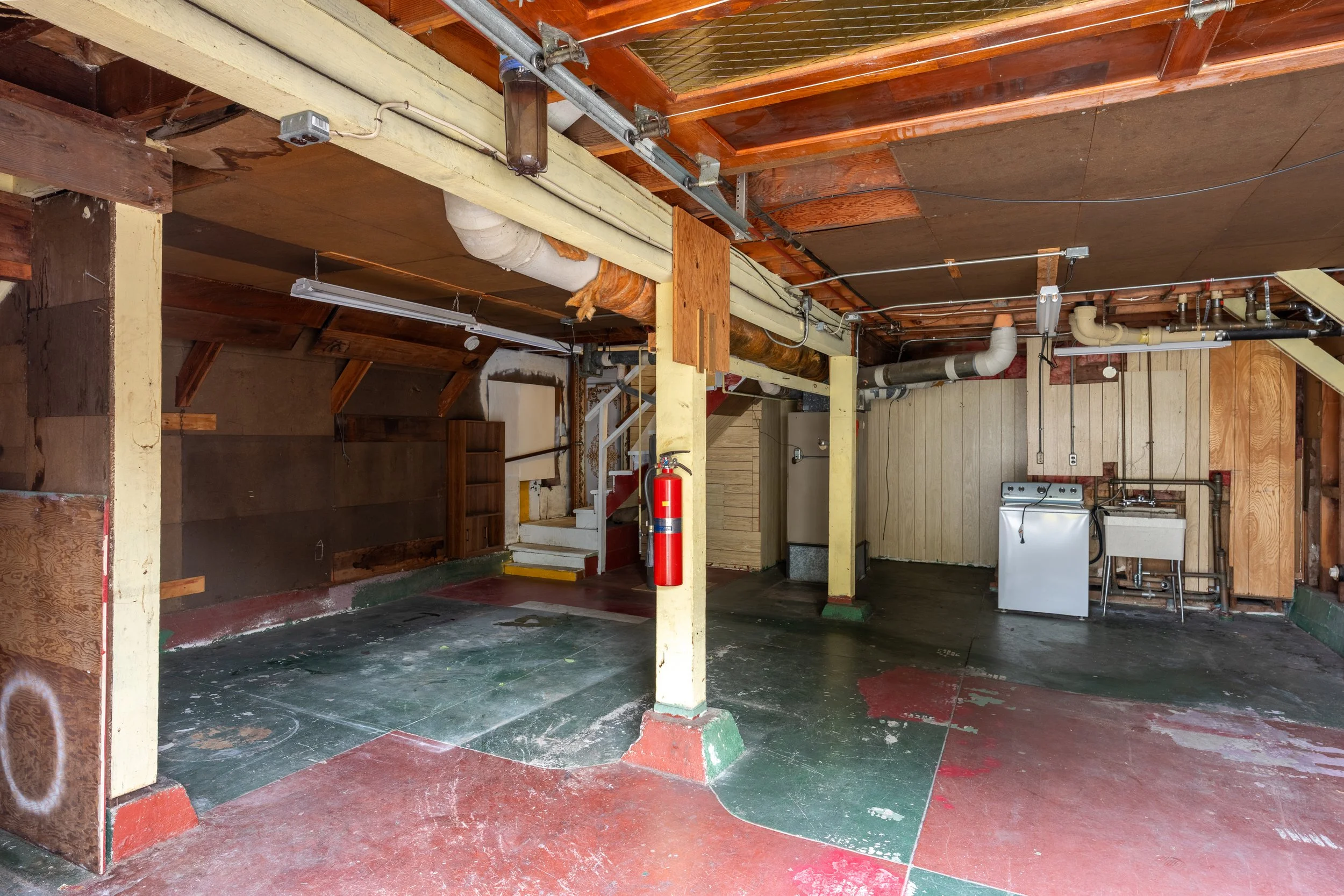 Interior view of a partially renovated garage or basement with exposed wooden beams, pipes, electrical wiring, and a laundry area in the back. There is a fire extinguisher on the floor near the center.
