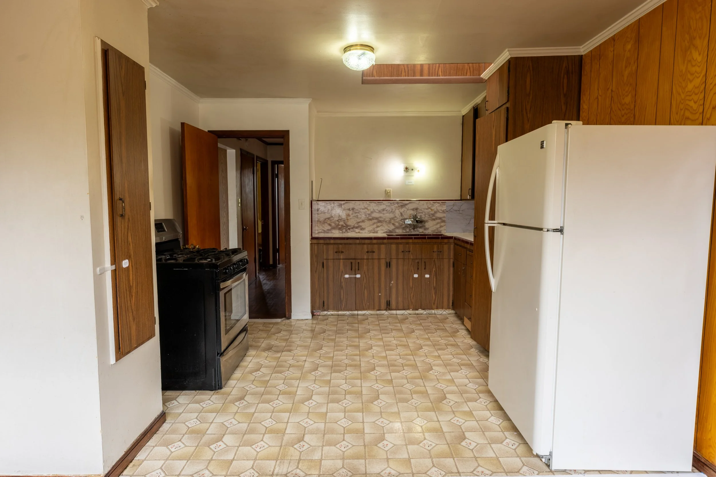 Kitchen with wooden cabinets, white refrigerator, black stove, and beige tile flooring, connected to hallway with wooden doors.