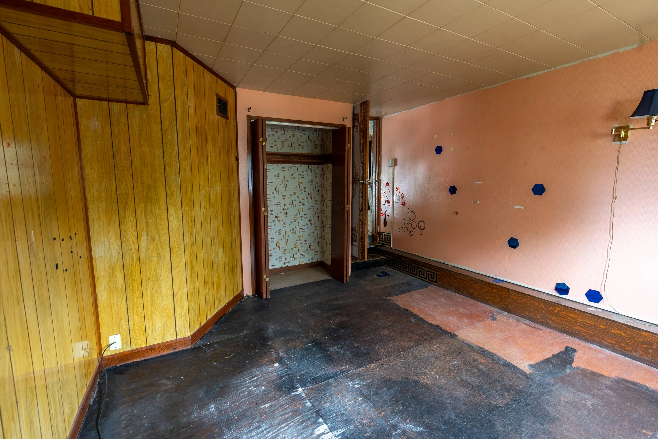 An interior room with wood-paneled walls, patterned wallpaper, and a partially removed black wooden floor. There is a doorway leading to another space with floral wallpaper, and a pink wall with blue hexagonal accents and a brass wall sconce.