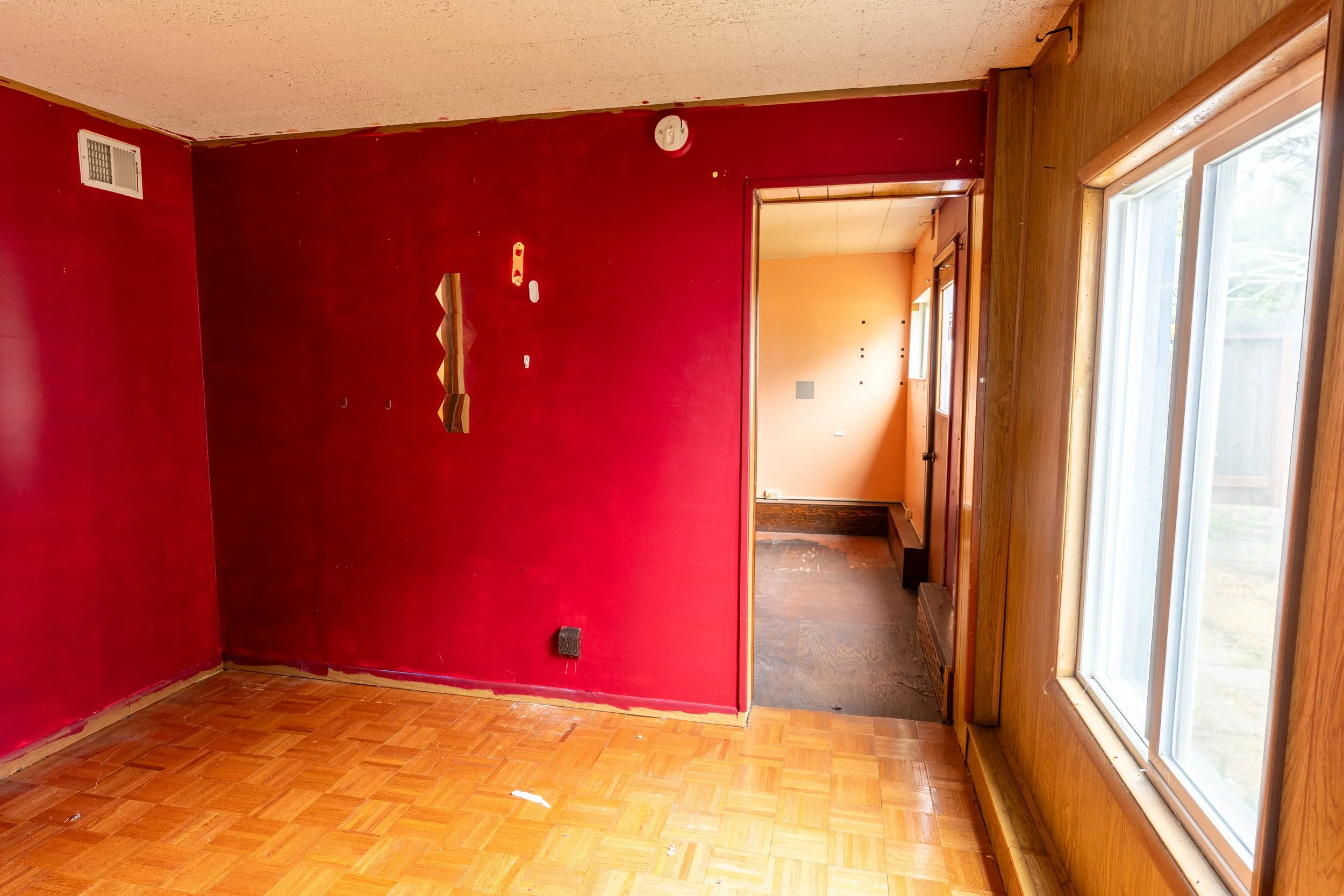 Empty room with red painted wall, wooden floor, and large windows, with some patches of paint removed and a doorway leading to another room.