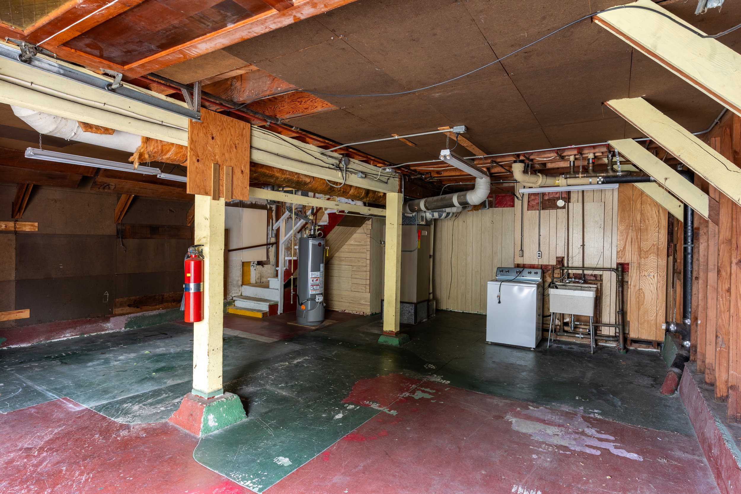 The image shows the unfinished interior of a building with exposed ceiling beams and pipes. The floor has patches of paint and concrete. A fire extinguisher is mounted on a yellow support column. There are octagonal wood and wood-paneled walls, with 