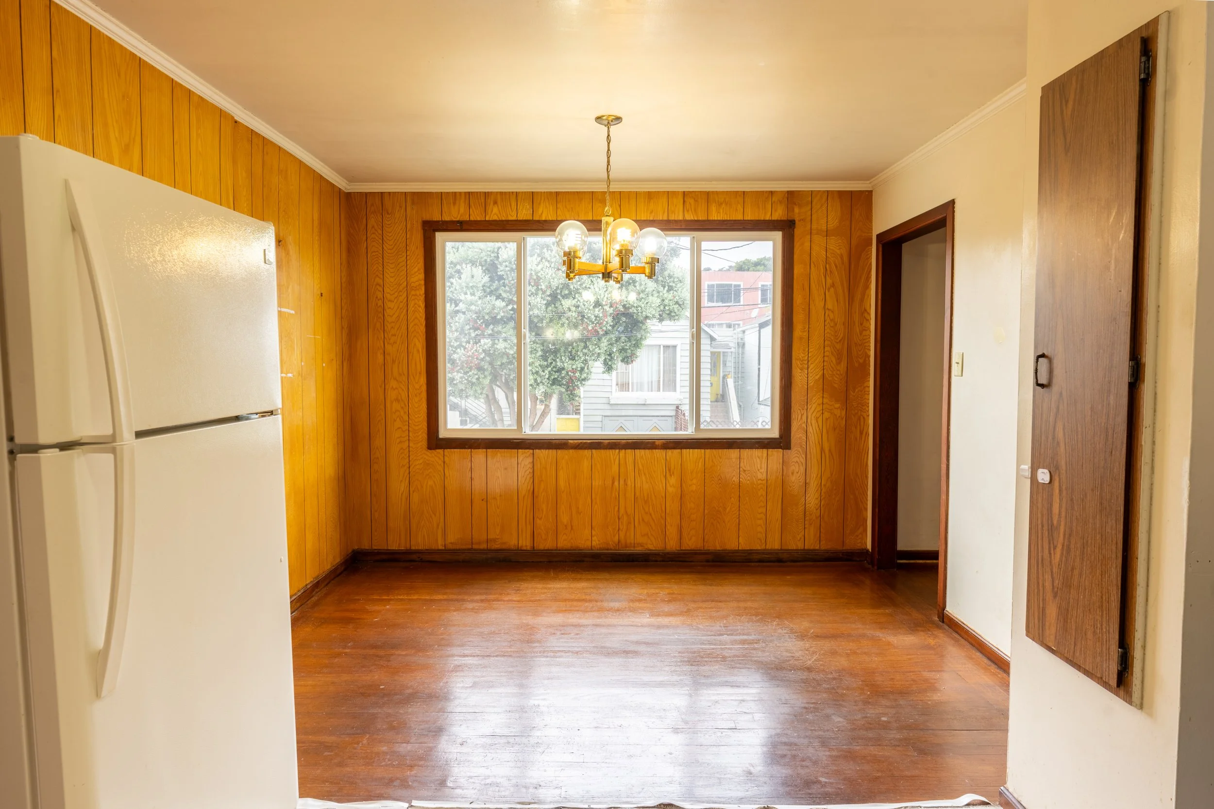 View of a room with wood-paneled walls, a hardwood floor, a large window showing trees and neighboring houses, a brass chandelier hanging from the ceiling, a white refrigerator partially visible on the left, and a door with a wooden frame on the righ
