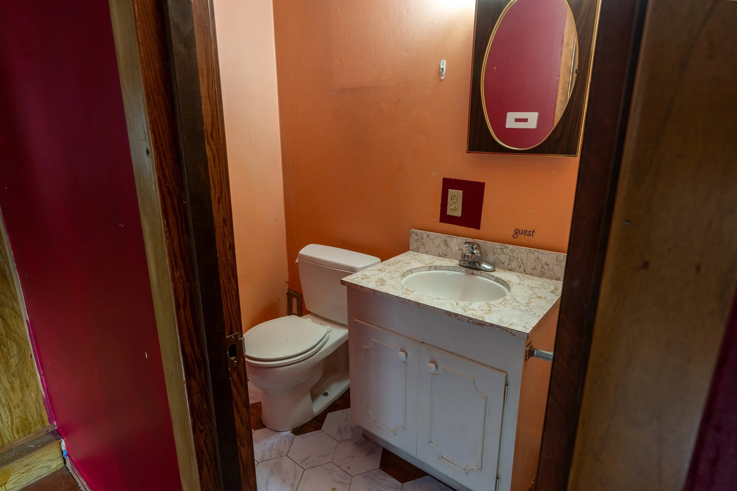 A small bathroom with an oval mirror above a marble countertop sink, a toilet, and an orange painted wall with a power outlet and a small hook. A wooden door frame is partially visible.