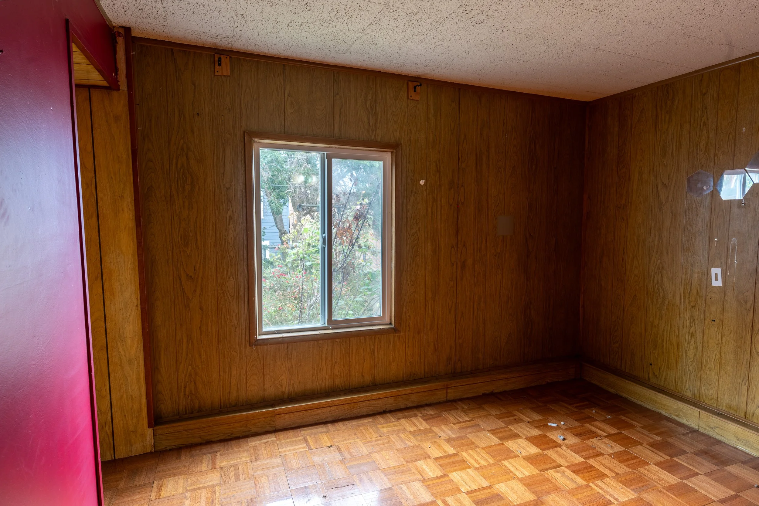 Empty room with hardwood floors, wood paneled walls, a window showing trees outside, and a pink wall on the left side.