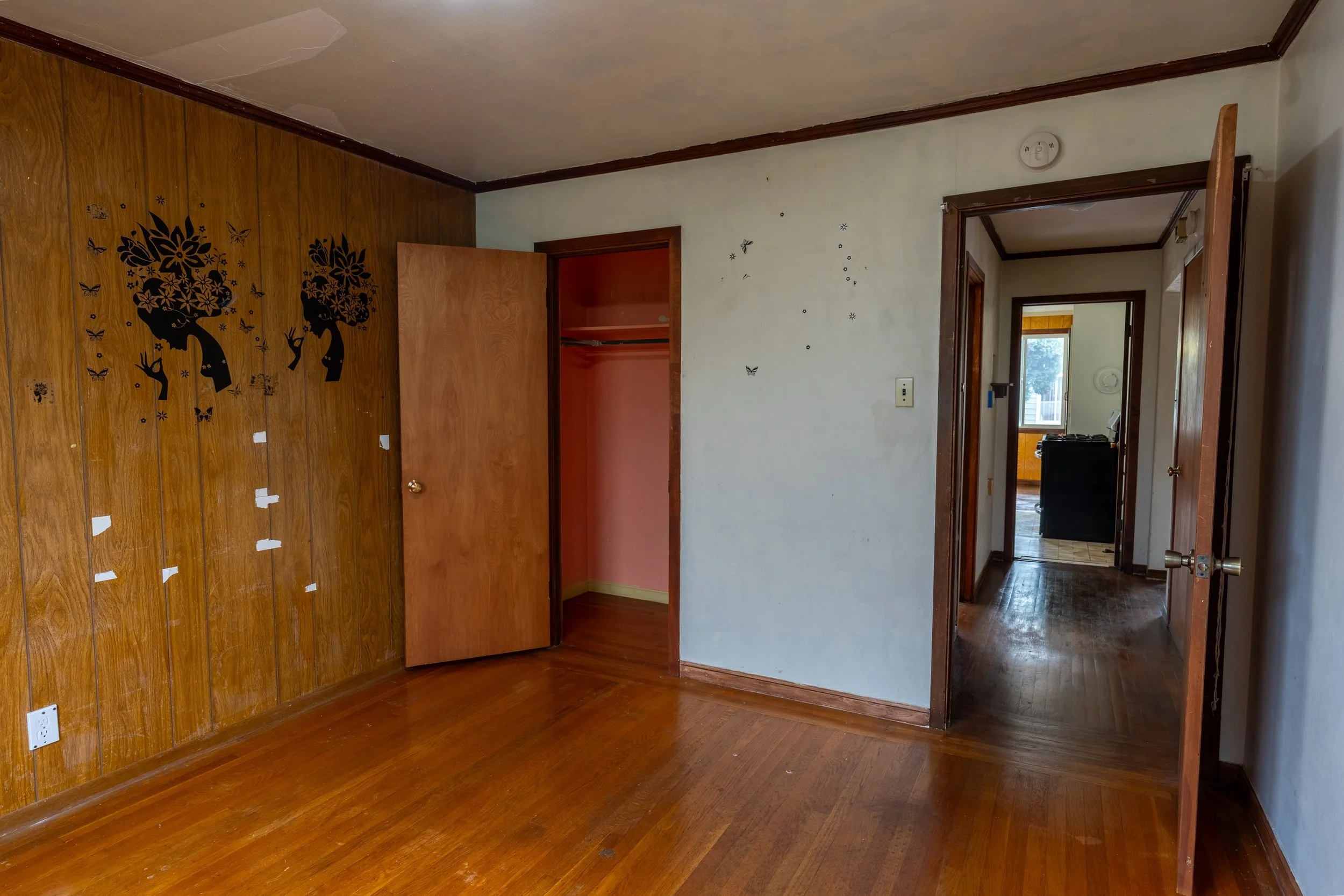 Empty room with wooden floors and wood-paneled walls, decorated with tree and butterfly wall decals, with open closet and view into hallway with windows.