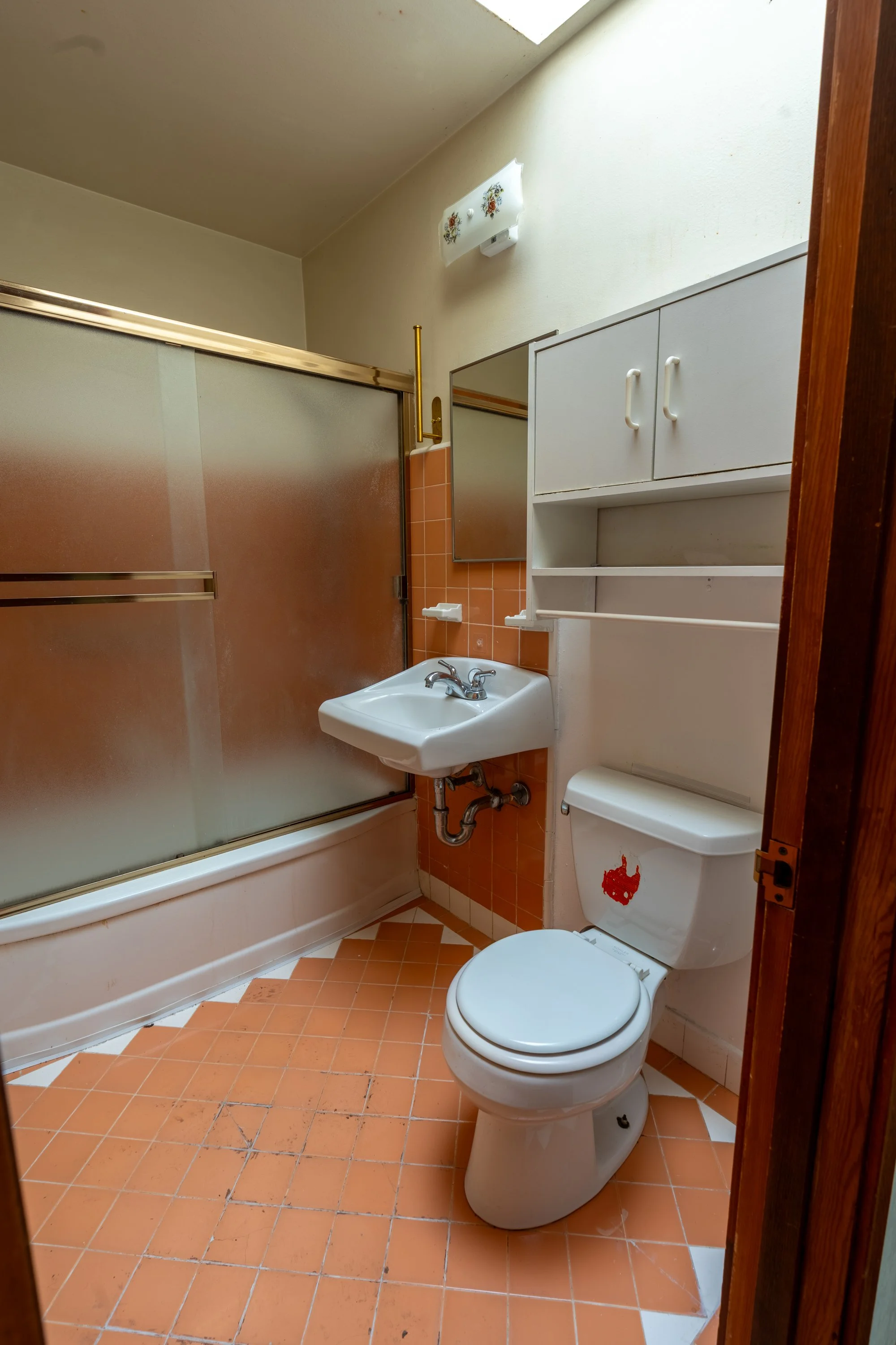 A small bathroom with a shower, sink, and toilet. The shower has frosted glass doors with a brass frame. The floor is tiled with orange tiles, and there is a white cabinet mounted above the toilet. The walls are painted beige, and there is a small wi