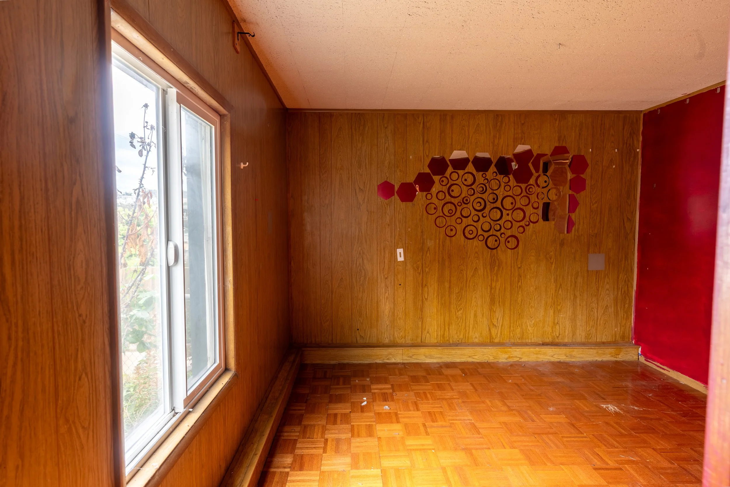 A room with wood-paneled walls, a large window on the left, and a decorative wall hanging composed of red hexagons and circles on the back wall. The floor is hardwood, and there is a red baseboard along the bottom of the walls.