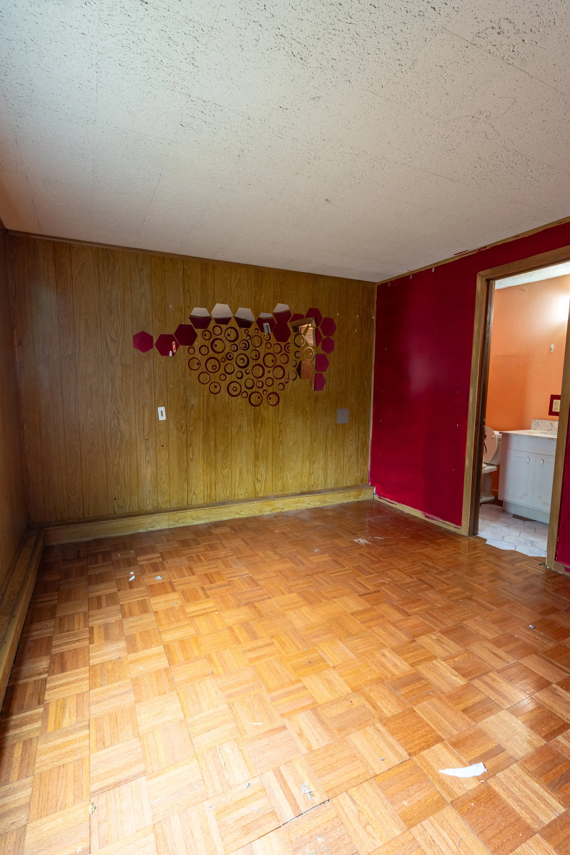 A room with wood paneling on two walls, red paint on one wall, parquet flooring, and a decorative wall piece made of red geometric shapes and circles. An open doorway reveals a bathroom with white cabinets and a washing machine.