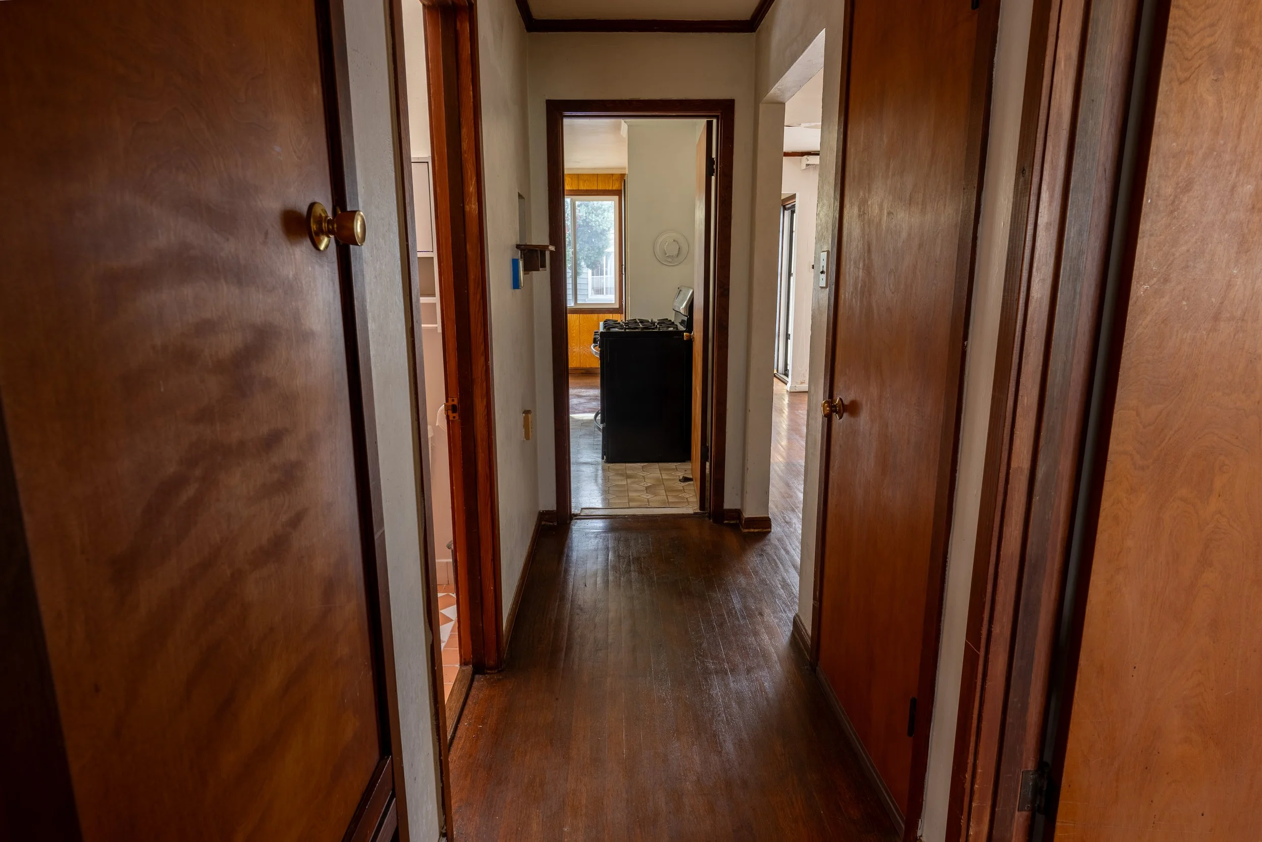 View through a hallway into a kitchen with a window, stove, and wooden paneling, with wooden doors on either side and hardwood flooring.
