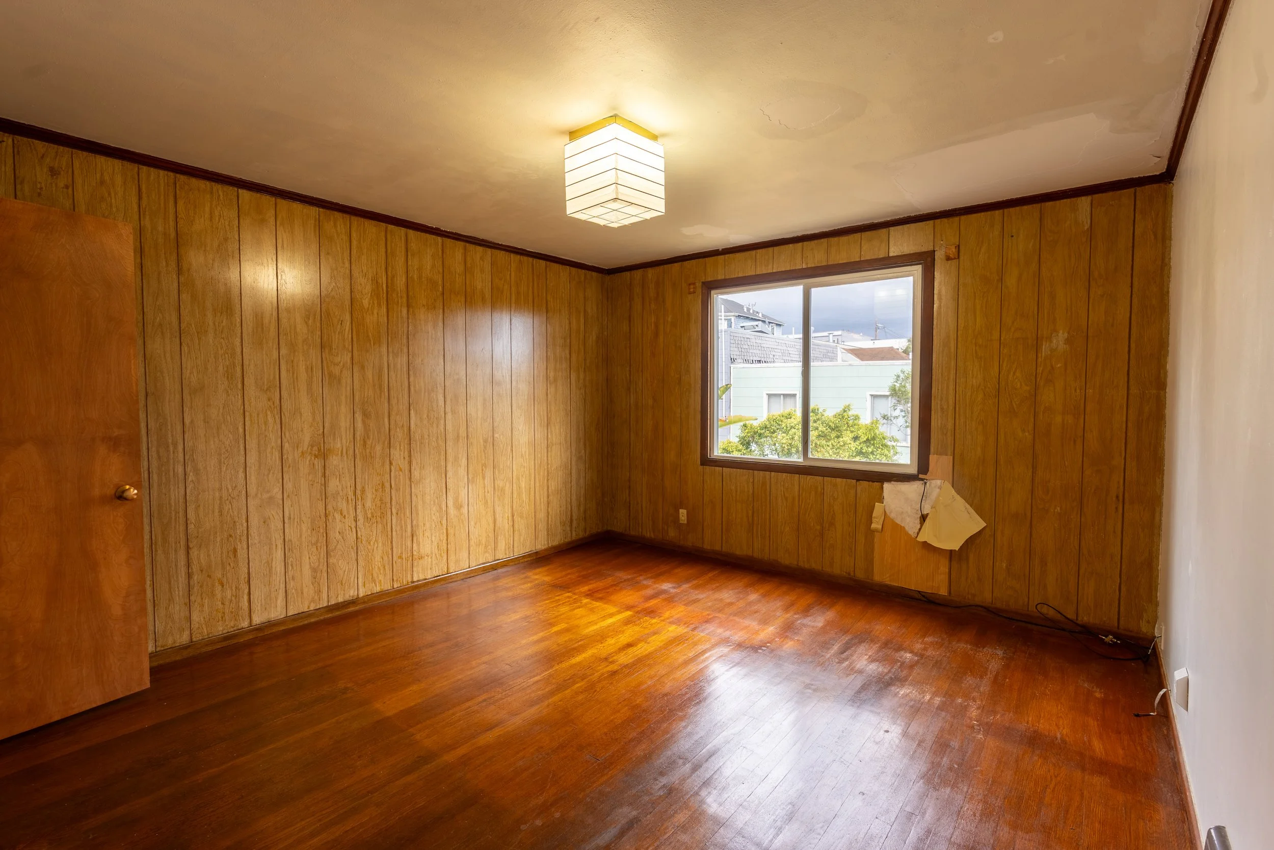 Empty room with wood-paneled walls, hardwood floor, a window showing neighboring houses, and a ceiling light fixture