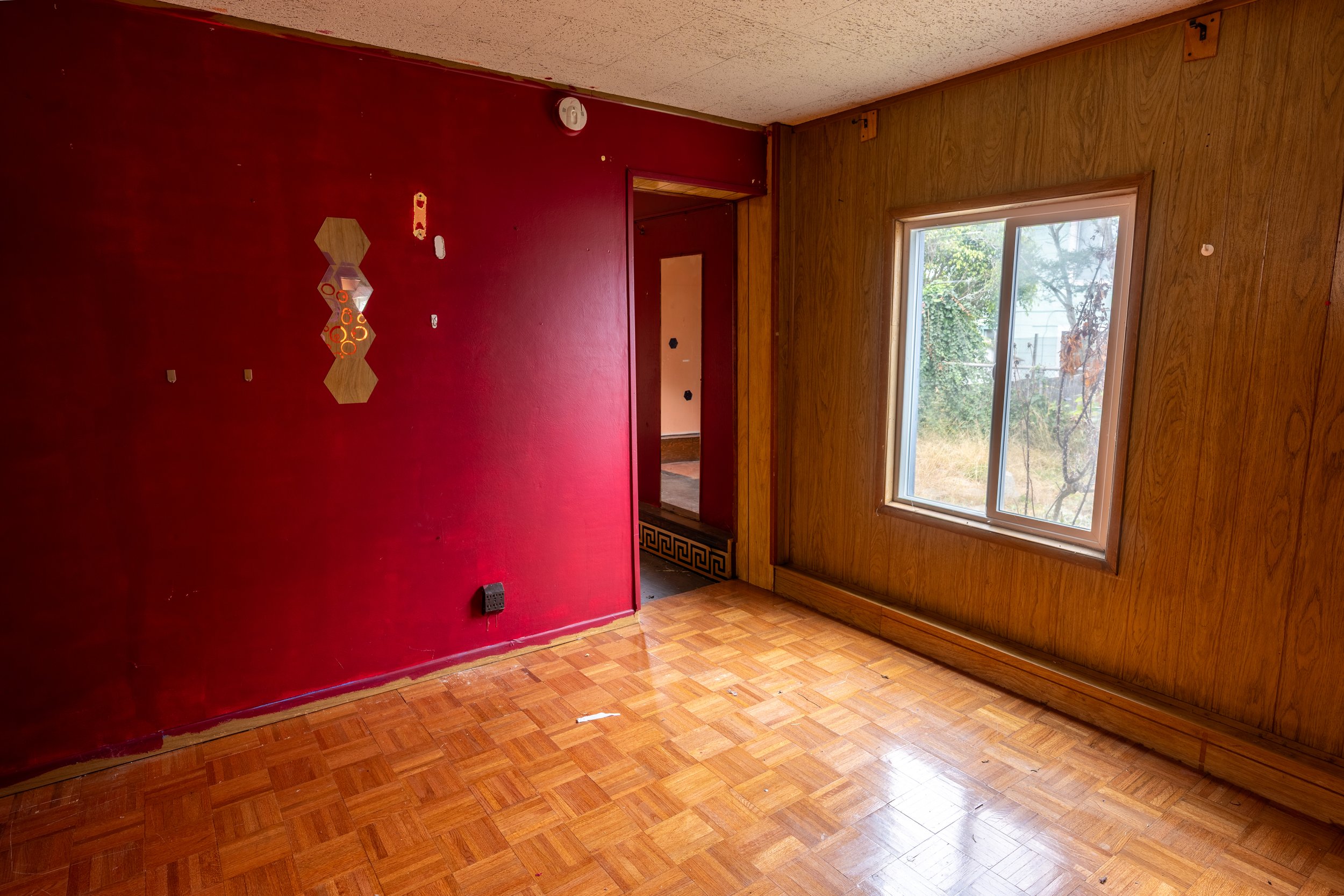 A room with wooden and red painted walls, a window showing trees outside, and a parquet floor. The red wall has some decorations, and there is a doorway leading to another room.