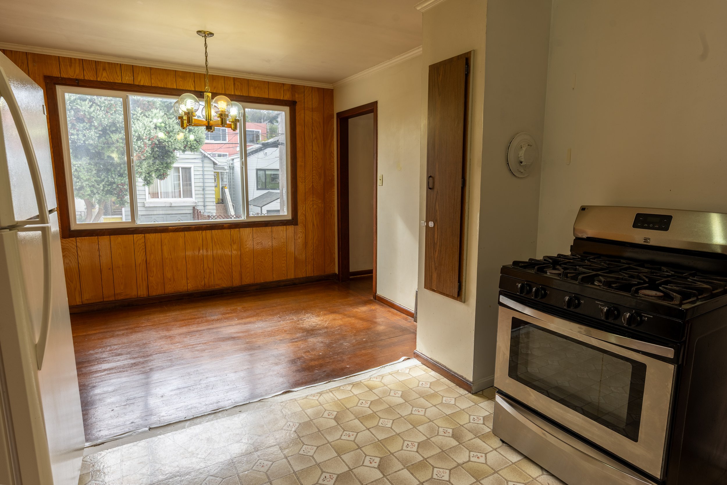 Interior of a kitchen with a wooden floor, large window, and a hanging light fixture with multiple bulbs. The kitchen has a gas stove on the right and a mix of wood-paneled and painted walls.