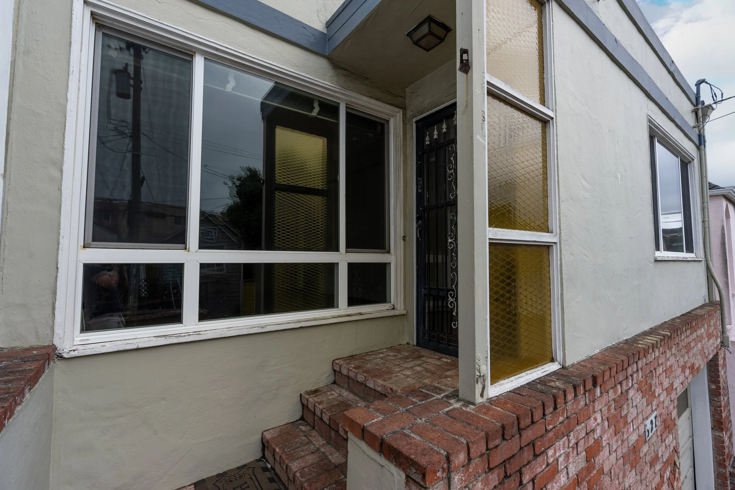 Front of a house with large windows and a red brick stoop leading to a black metal door. The house exterior is painted light yellow with decorative yellow textured panels and a decorative black light fixture above the entrance.