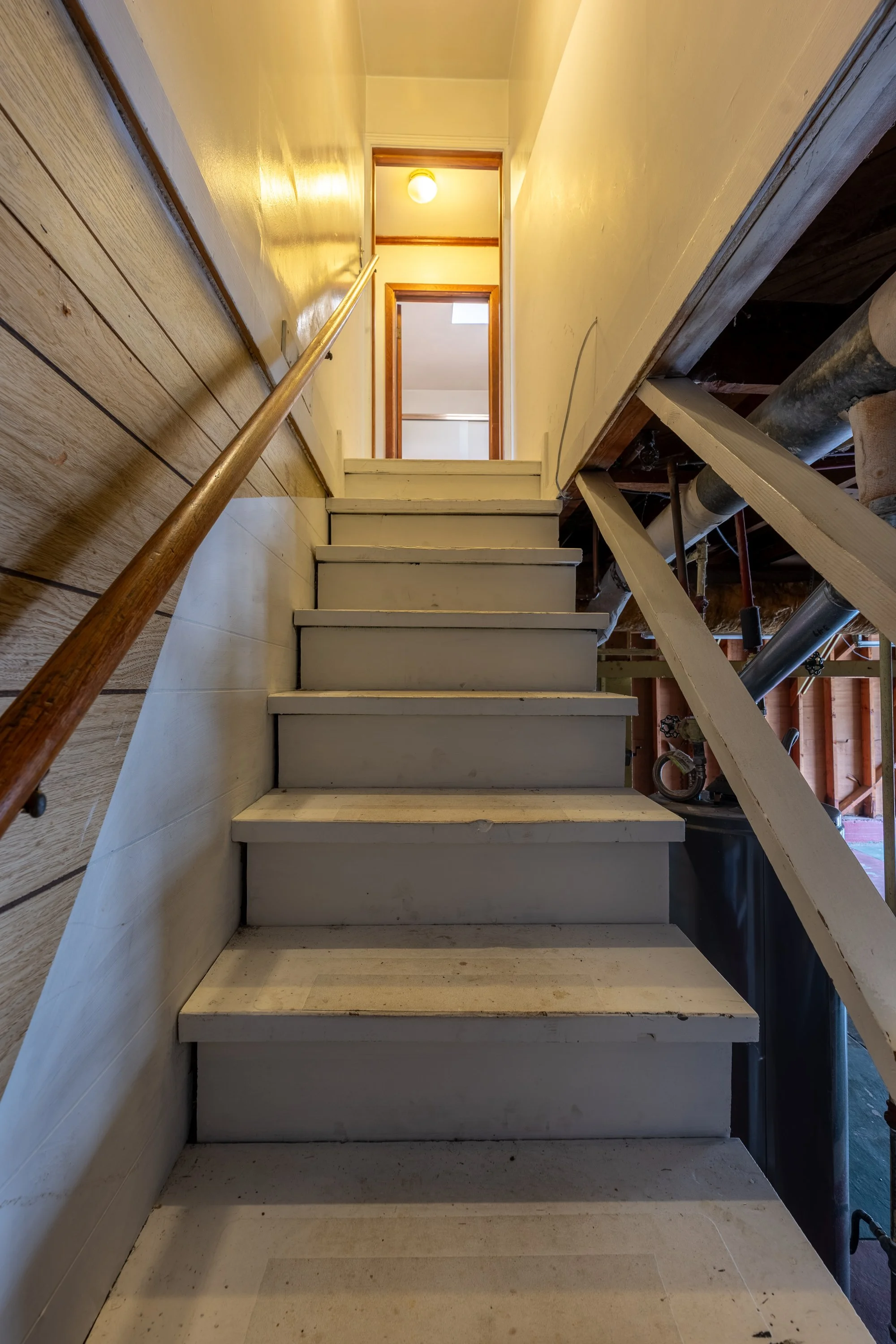 View of a partially built staircase with white unfinished steps, a wooden handrail on the left, and exposed pipes and beams on the right, with a yellow ceiling light overhead.