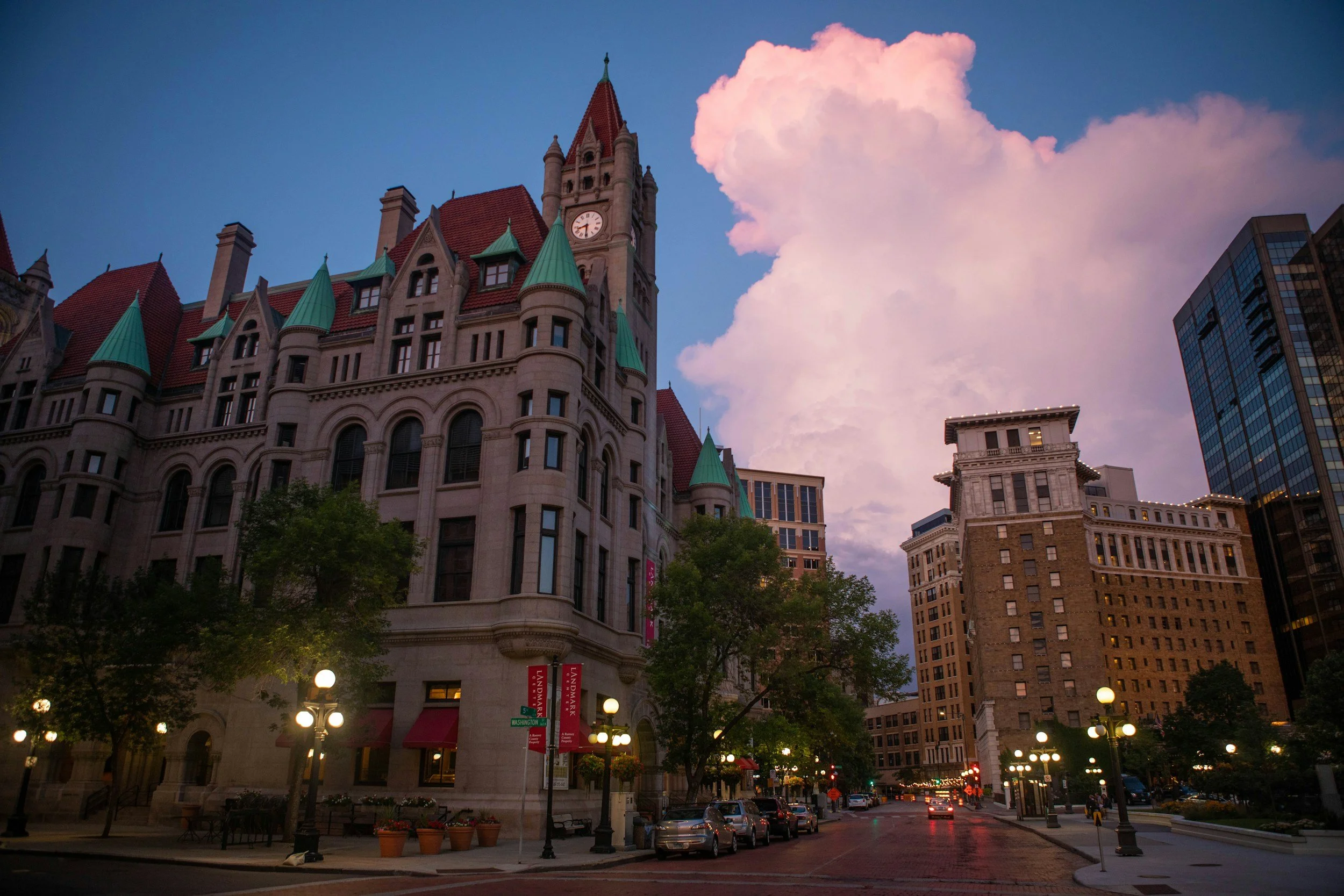 A historic building with green rooftops and a clock tower, surrounded by modern glass buildings and trees, under a sky with pink clouds.