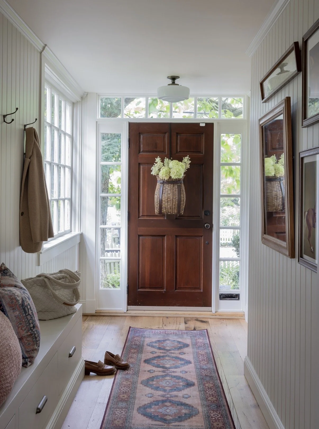 Entryway with a wooden front door, white walls, and glass panels on either side. A hanging plant in a woven basket decorates the door. To the left, a coat is hanging on a hook near a window with a bench below, holding a quilted bag and shoes. To the right, framed artwork hangs on the wall. A small rug runs from the door into the room.
