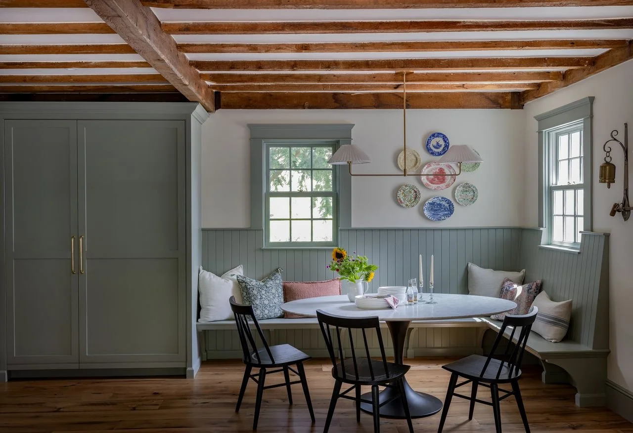A cozy dining nook with a round table, black chairs, and built-in bench with cushions, set against white and sage green walls with wooden ceiling beams.