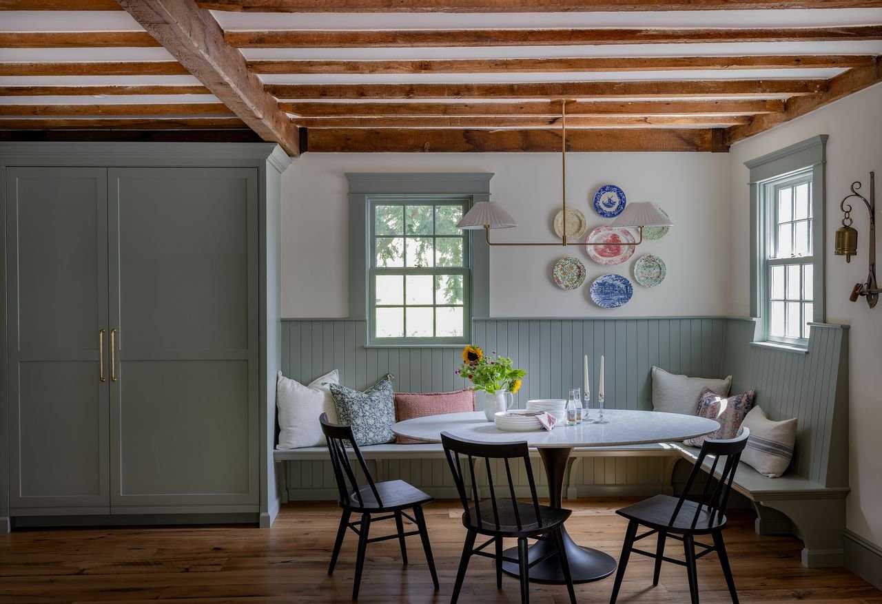 A cozy dining nook with a round table, black chairs, and a built-in bench with cushions under two windows. The wall behind the bench has decorative plates, and the ceiling has exposed wooden beams.