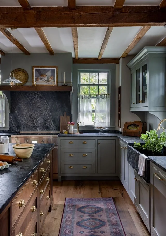 Cozy kitchen with wooden beams on ceiling, green cabinets, black countertops, window with white curtains, and a colorful rug on wooden floor.