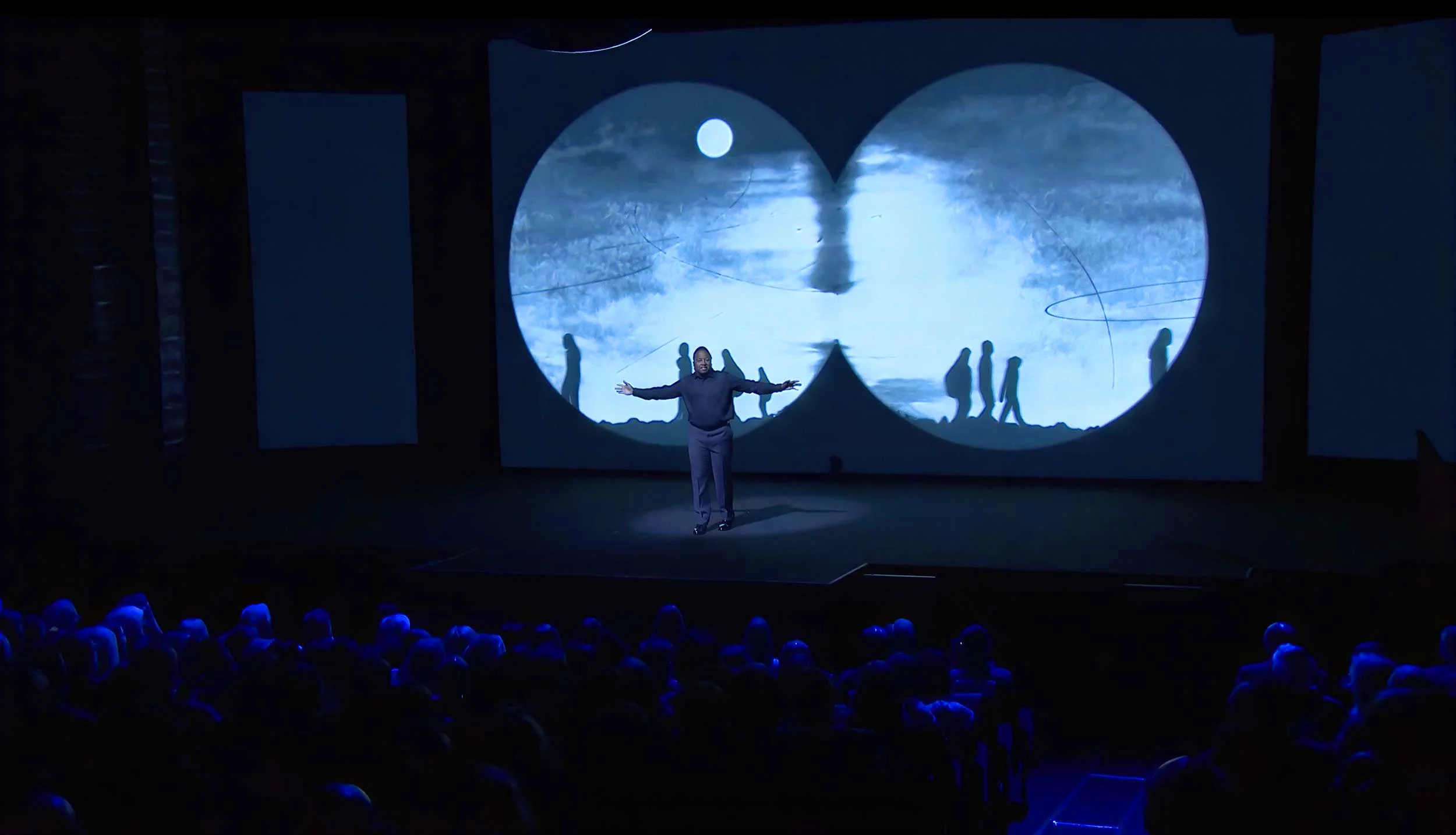 On stage at the Skoll World Forum in Oxford, Darius Simpson spreads his arms before two projected circles of moonlit landscape — The Power of Proximity, Skoll World Forum.
