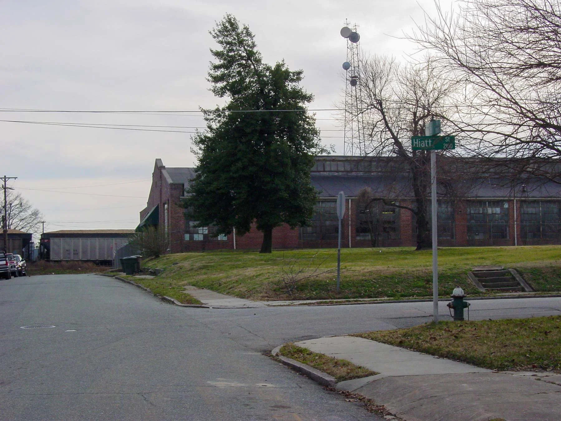 A quiet street corner by a brick industrial building, near where the house once stood — The Renter.