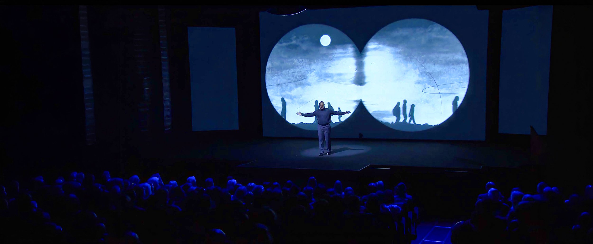 Darius Simpson performs with arms open as silhouetted figures walk across a binocular-shaped projection — The Power of Proximity, Skoll World Forum.