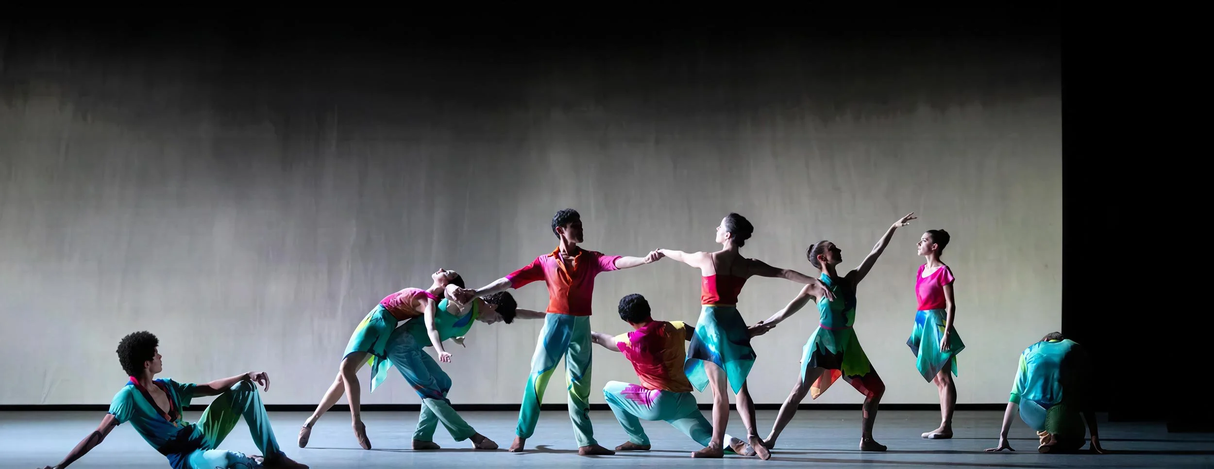 Dancers in painted costumes of teal, magenta and orange link hands across the stage in front of a giant screen of projected painted rain — Petrichor, Miami City Ballet.