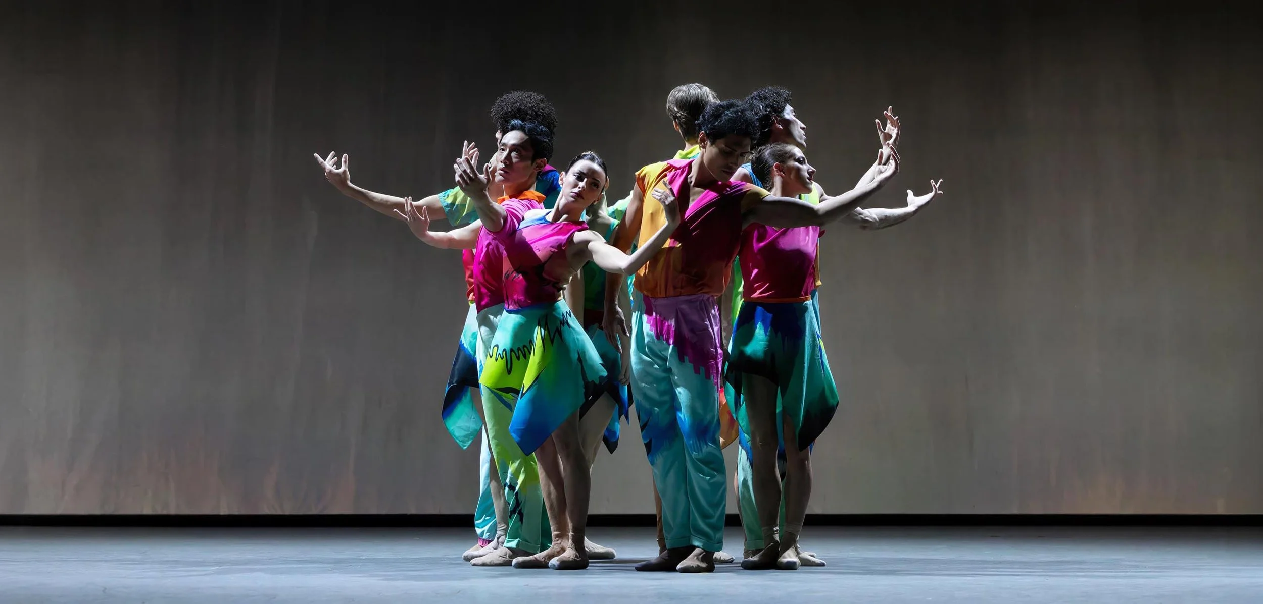 A cluster of dancers in teal and magenta costumes reach outward in front of pale projected rain — Petrichor, Miami City Ballet.