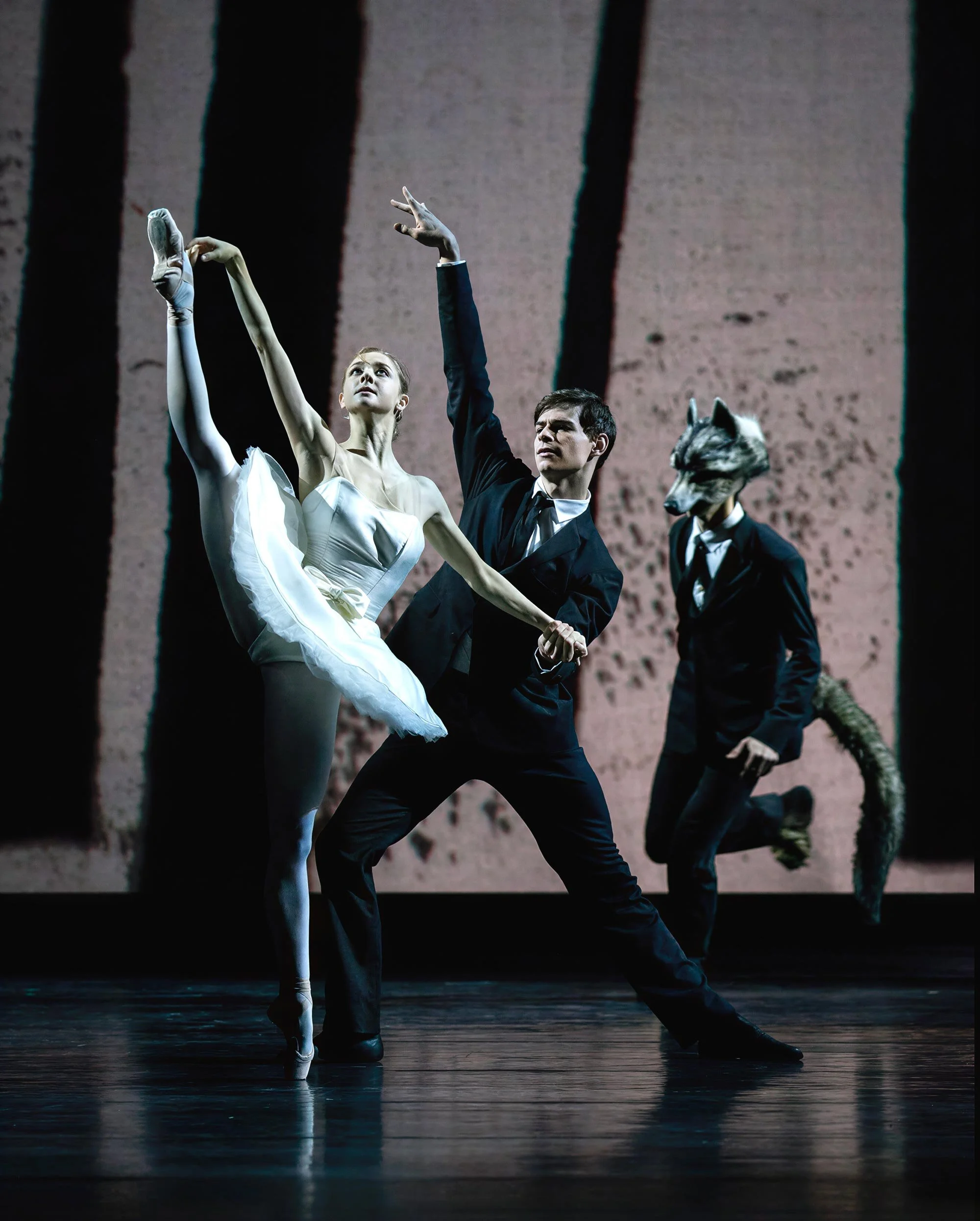 A ballerina in a white tutu extends her leg high beside a dancer in a dark suit, as wolf-masked figures move through the projected trees — Between Dogs & Wolves, Wiener Staatsballett.