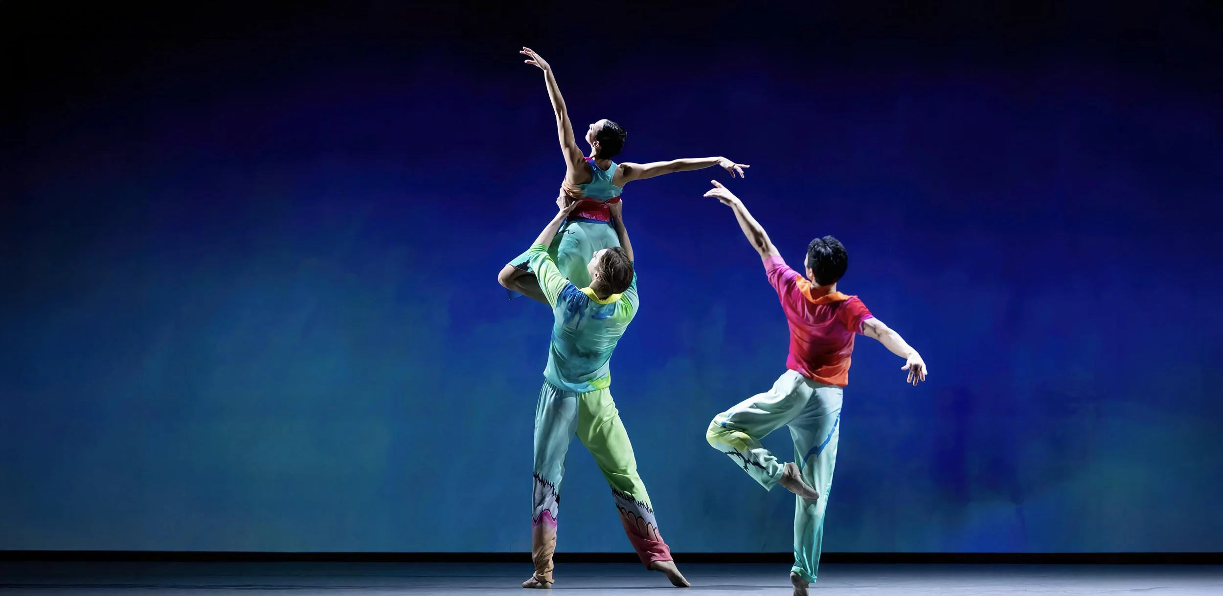 In colorful costumes, a male dancer lifts a ballerina in front of a projected painting of rain — Petrichor.