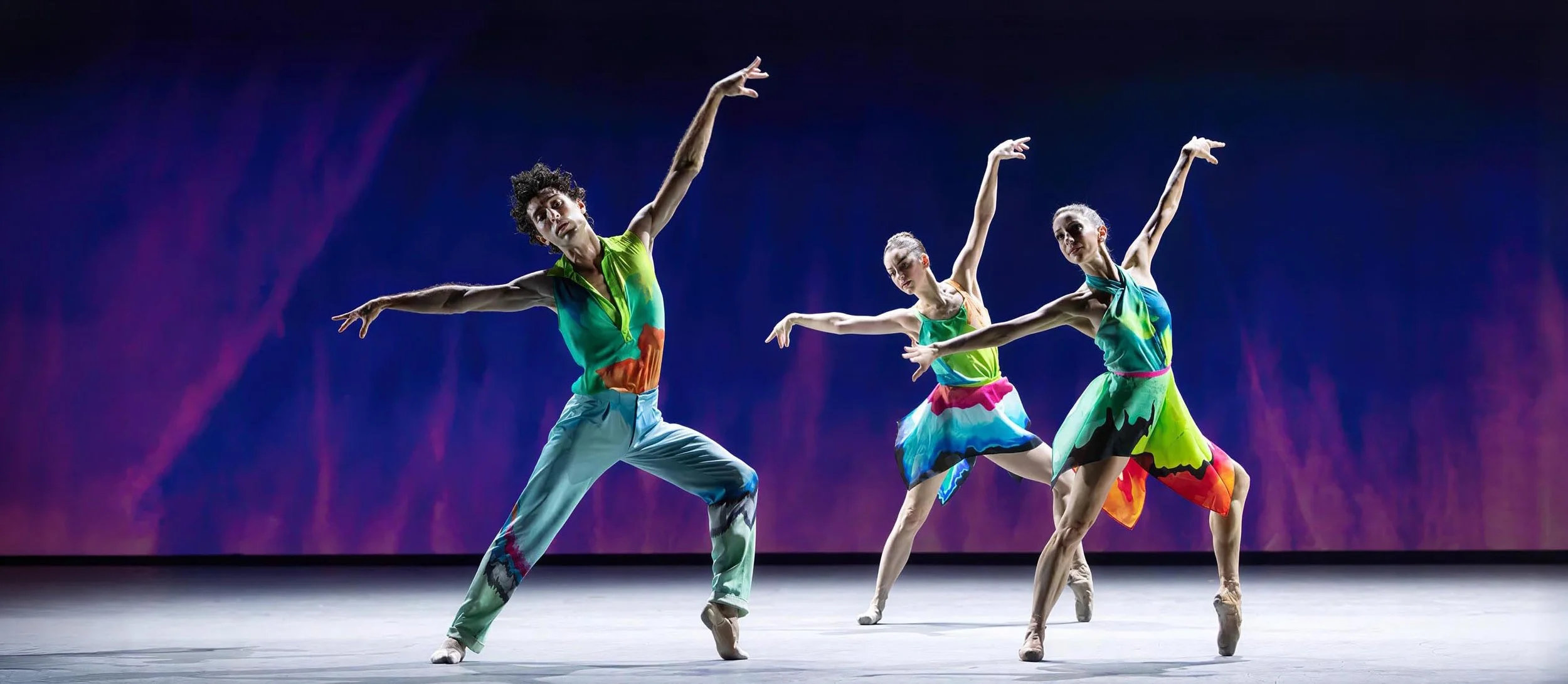 Three dancers leap in painted costumes against projected rain glowing violet and deep blue — Petrichor, Miami City Ballet.