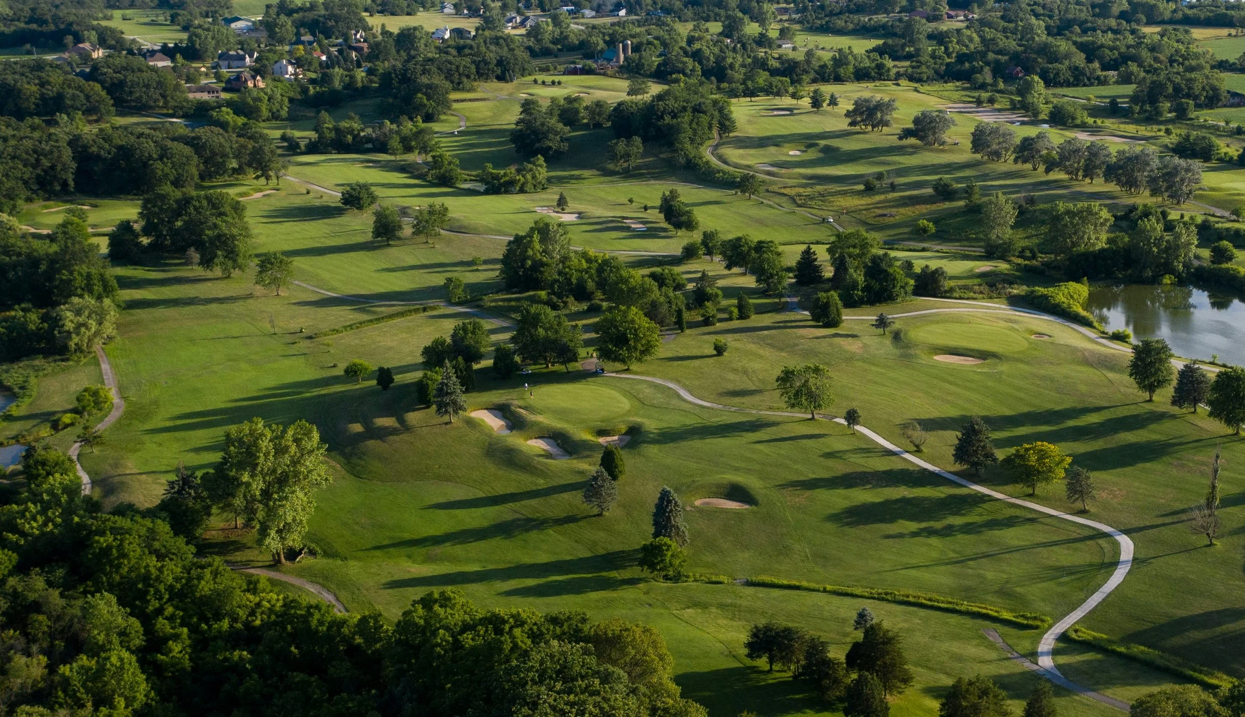 Aerial view of a golf course with green fairways, sand bunkers, and scattered trees, surrounded by a residential area and fields.