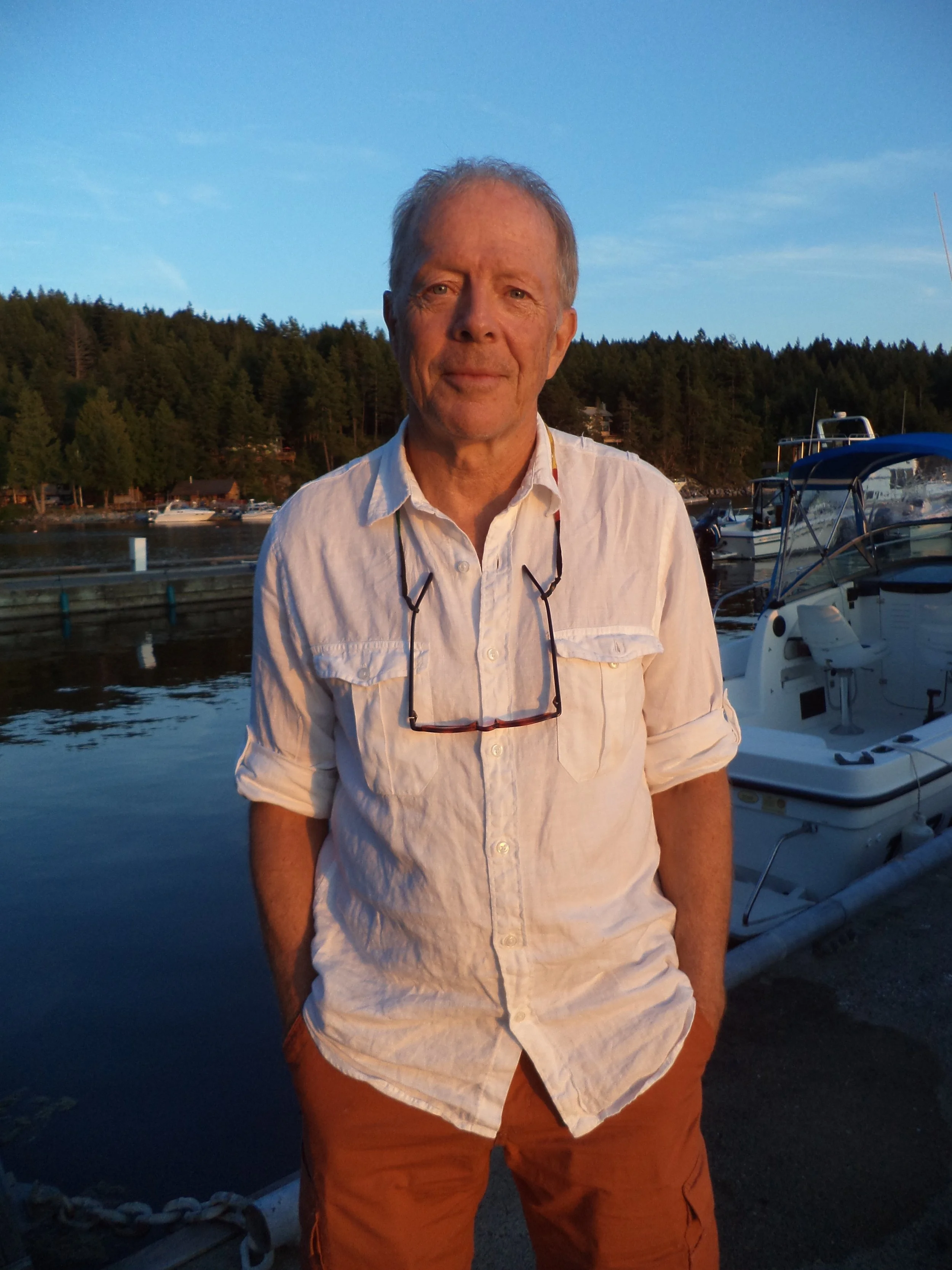 A middle-aged man with light skin and short light hair, wearing a light-colored button-up shirt with sunglasses hanging around his neck, stands in front of a marina with boats and a forested hillside in the background during sunset.