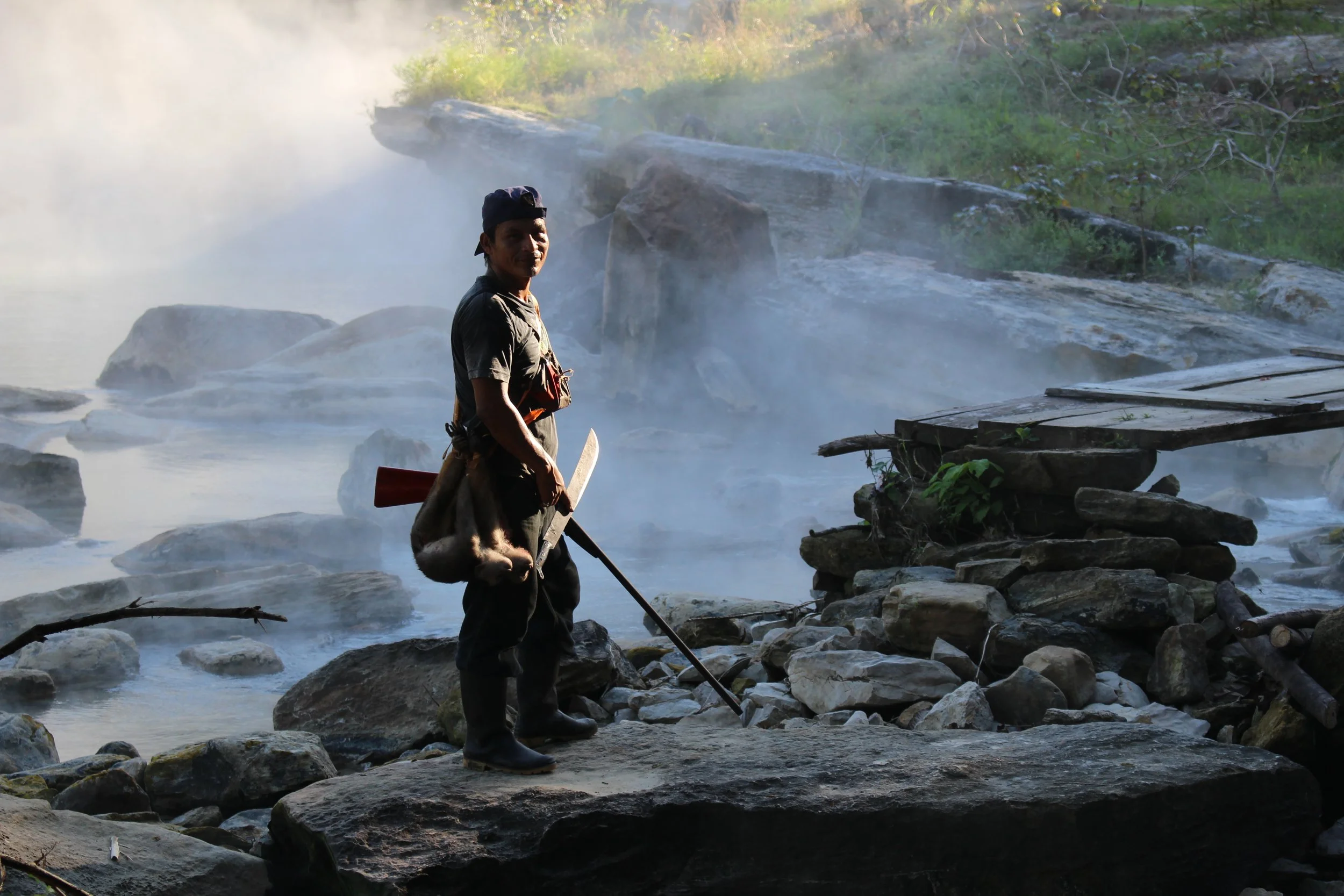 A person standing on rocks near a hot spring or geyser, with steam rising and lush greenery in the background.