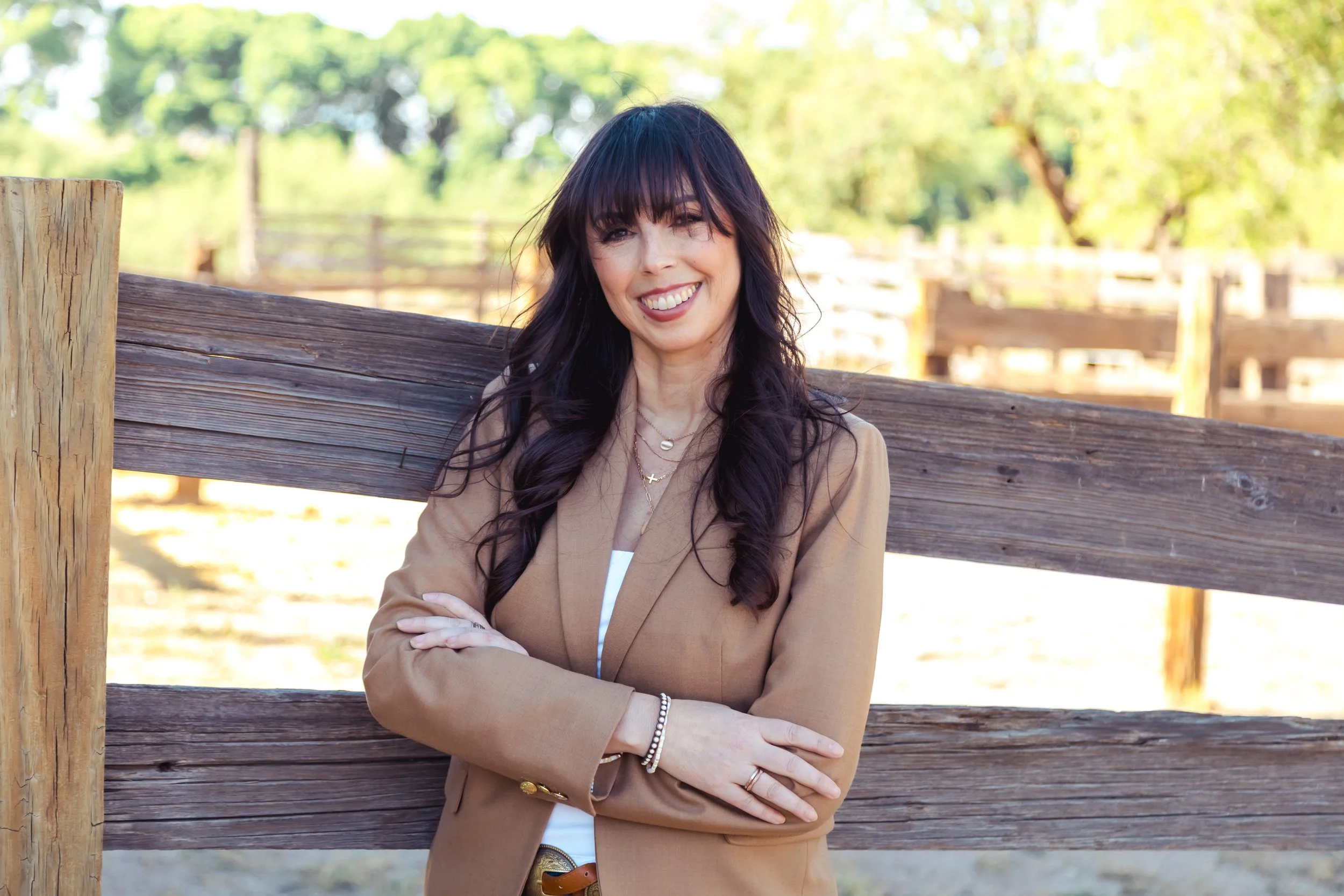 A woman with long dark hair and bangs, smiling, standing outdoors near a wooden fence with green trees in the background.
