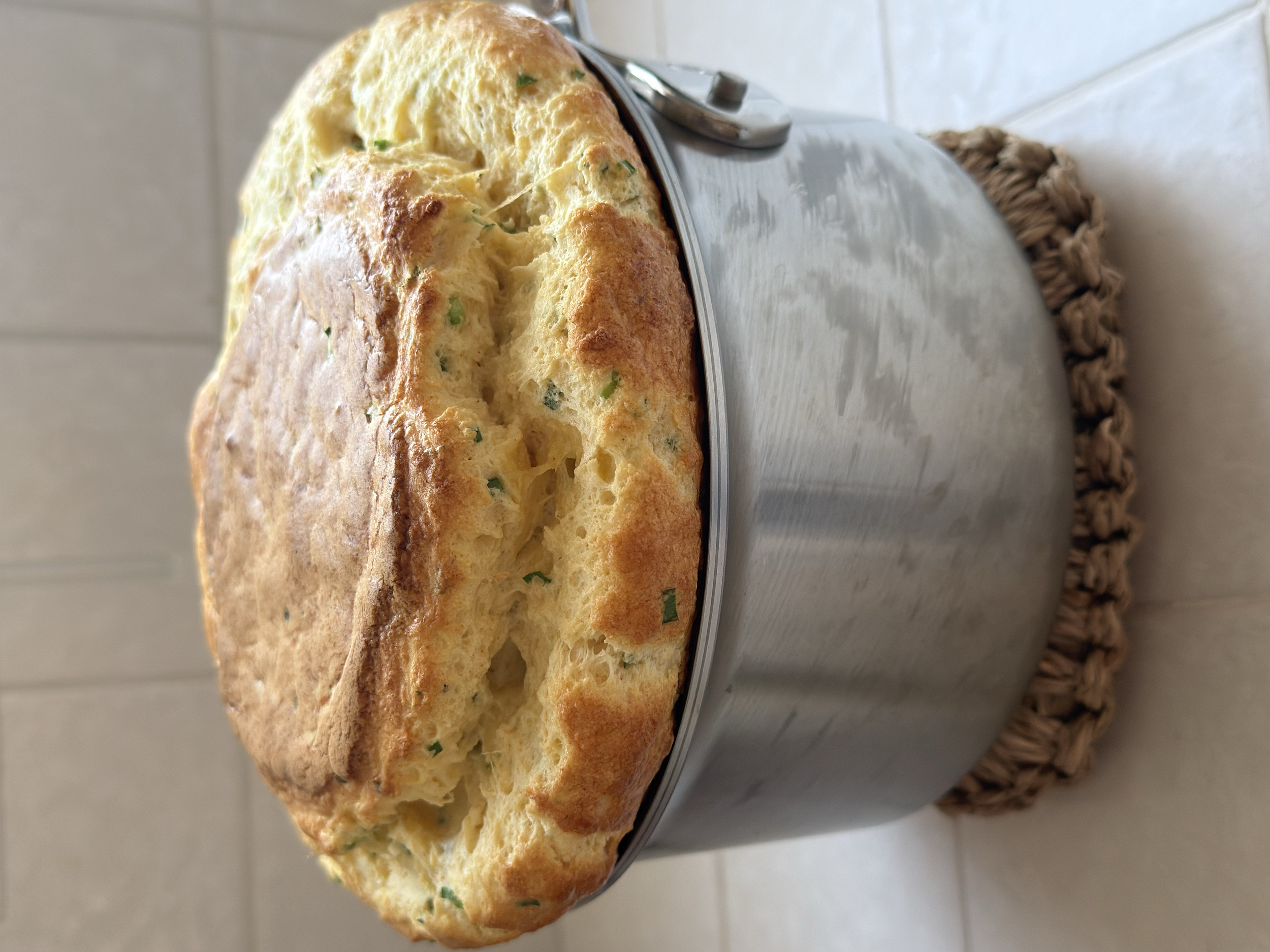 Close-up of a fluffy, baked bread or pastry with green herbs on top, sitting in a metal bowl placed on a woven mat.
