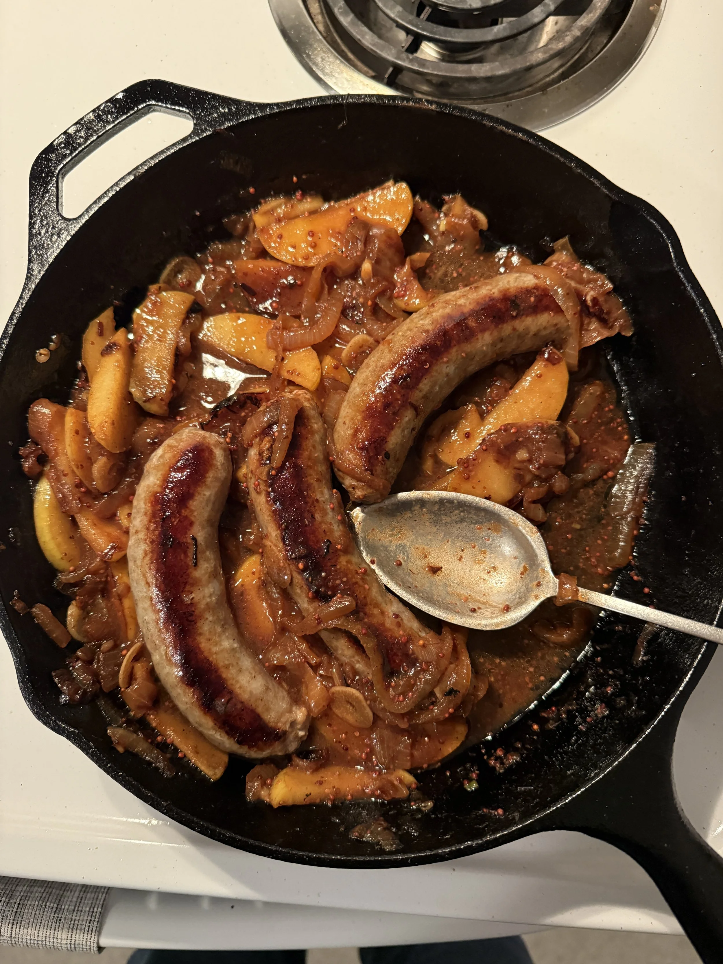 Cooking skillet containing grilled sausages, yellow and orange bell peppers, onions, and spices on a stove.