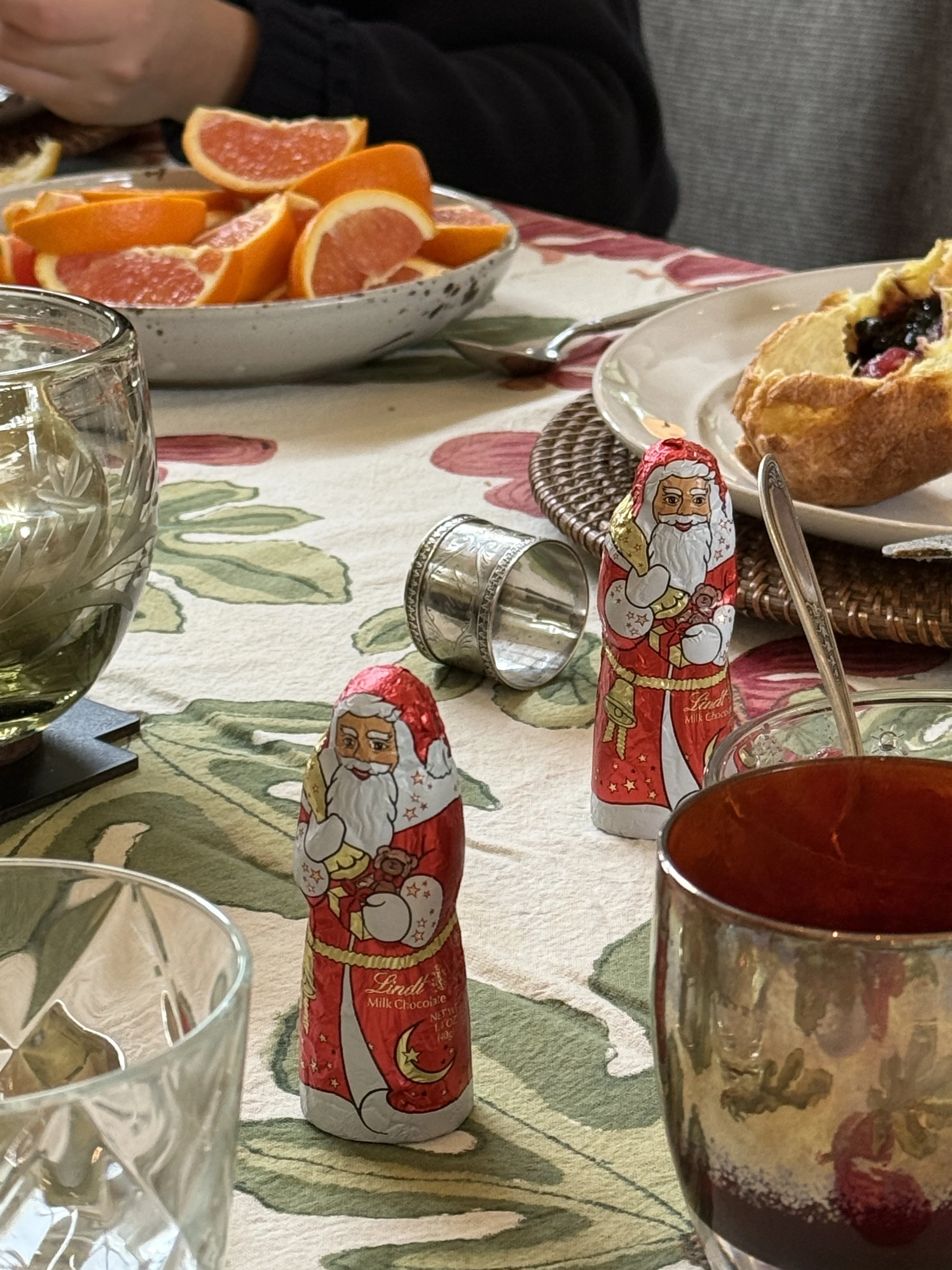 A table with a bowl of sliced grapefruits and oranges, a plate with a blueberry pastry, glasses of water, a candy with Santa Claus and a tin container.
