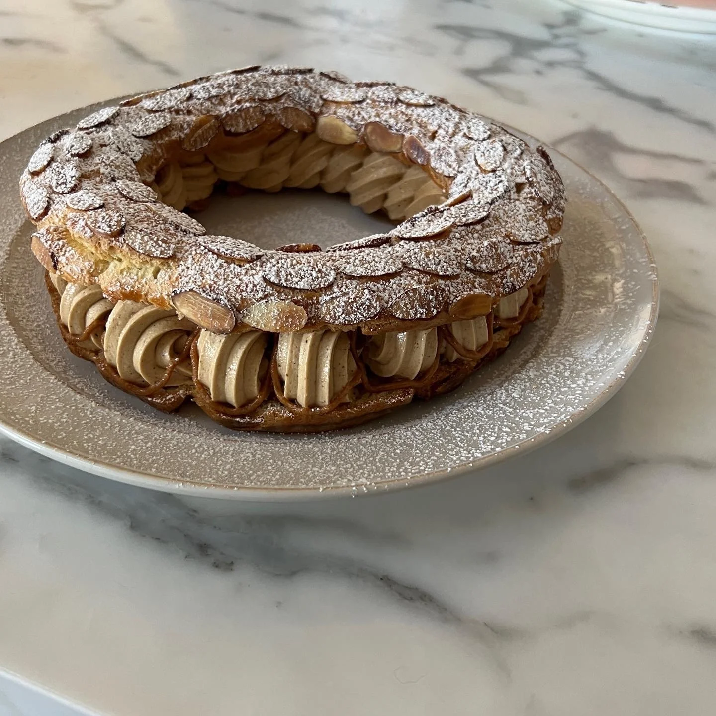 A decorative ring-shaped pastry with almond slices and a dusting of powdered sugar, filled with piped caramel-colored cream on a silver plate.