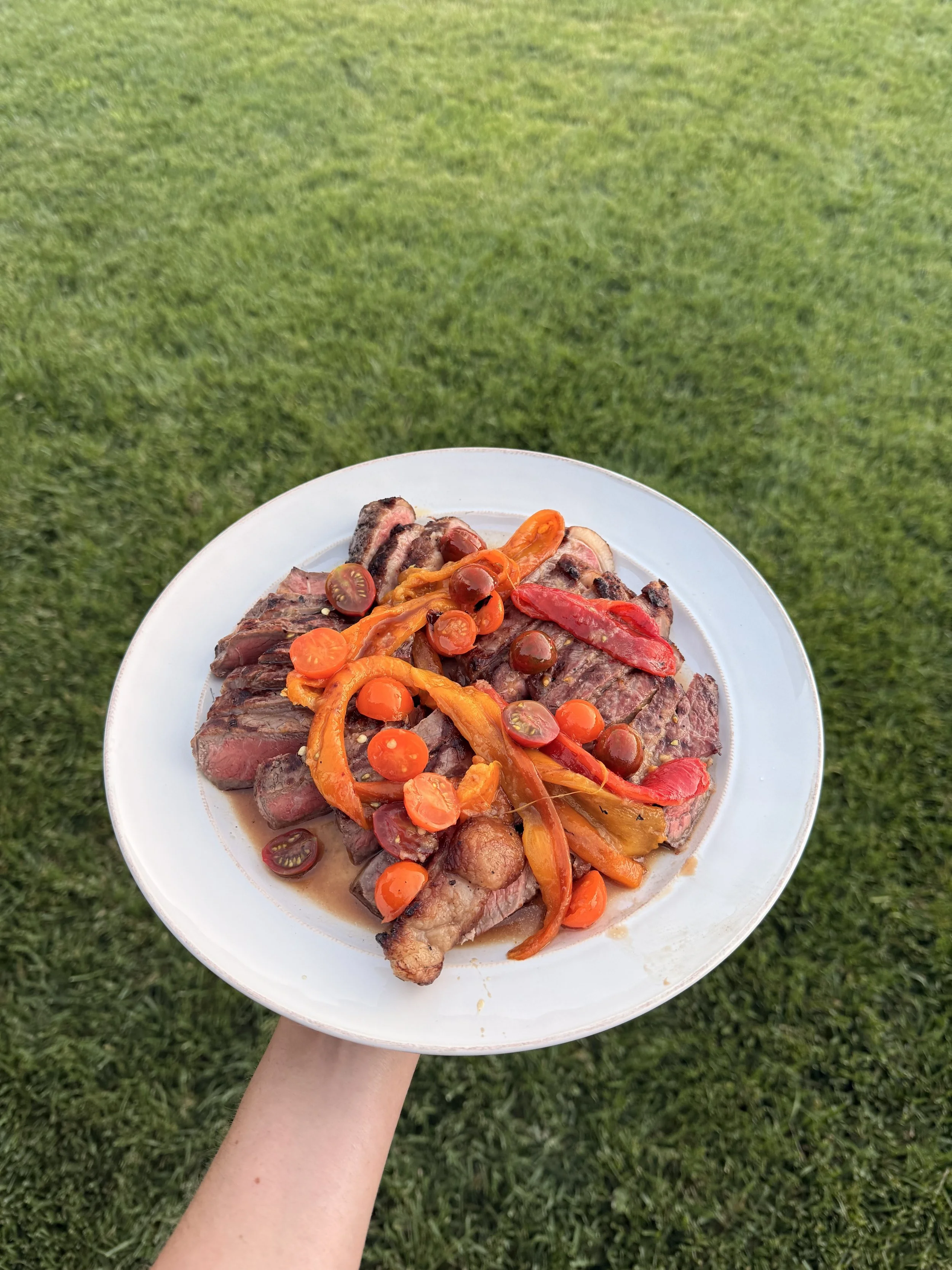 A person holding a white plate with cooked steak slices topped with colorful roasted peppers and cherry tomatoes on a grassy background.