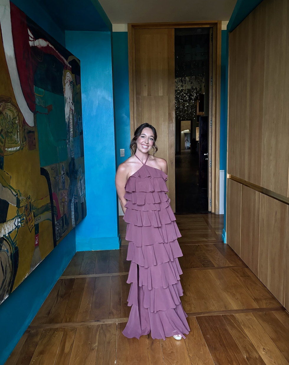 A woman in a pink ruffled dress standing inside a room with wooden floors, a colorful abstract painting on the wall, and a doorway leading to a dimly lit hallway.