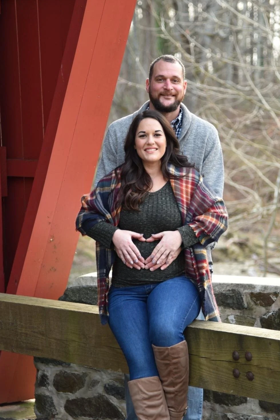 A couple standing outdoors with the woman sitting on a wooden railing and the man standing behind her. The woman has dark hair, is wearing a plaid jacket, dark top, blue jeans, and brown boots. They are smiling and holding their hands over her pregna